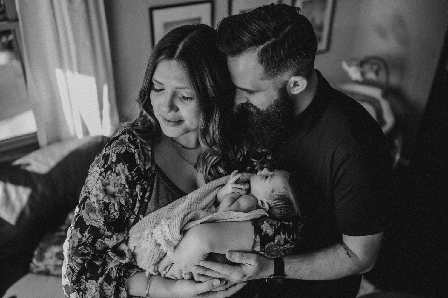 Black and white portrait of family bonding with newborn in sunlit room.