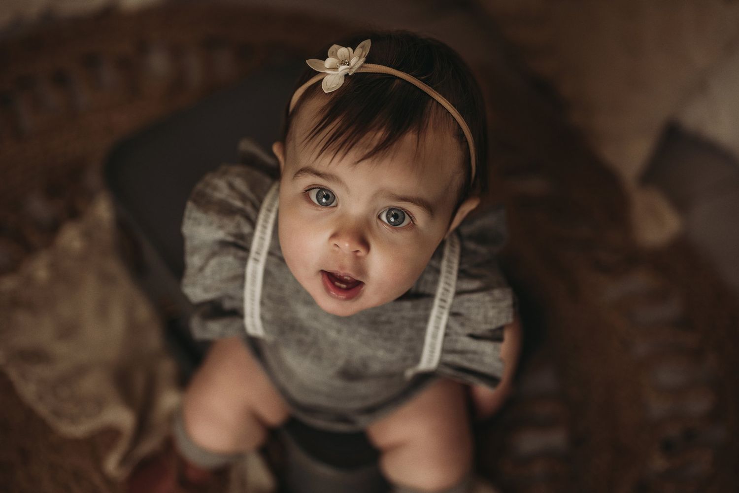 A small child wearing a gray ruffled romper and delicate headband looking upward with an excited expression.