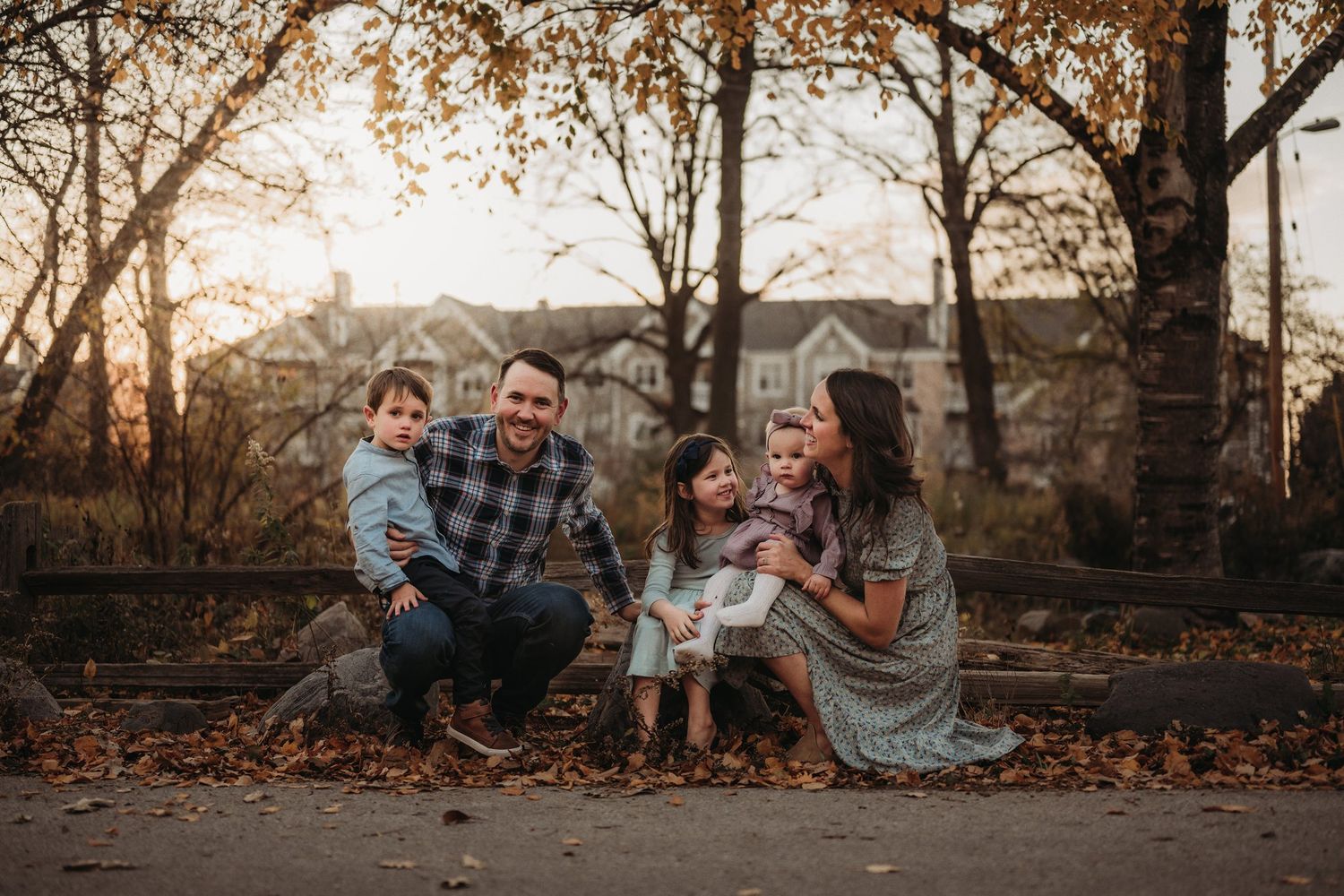 A family sits together on fallen autumn leaves at sunset with suburban homes in the background.