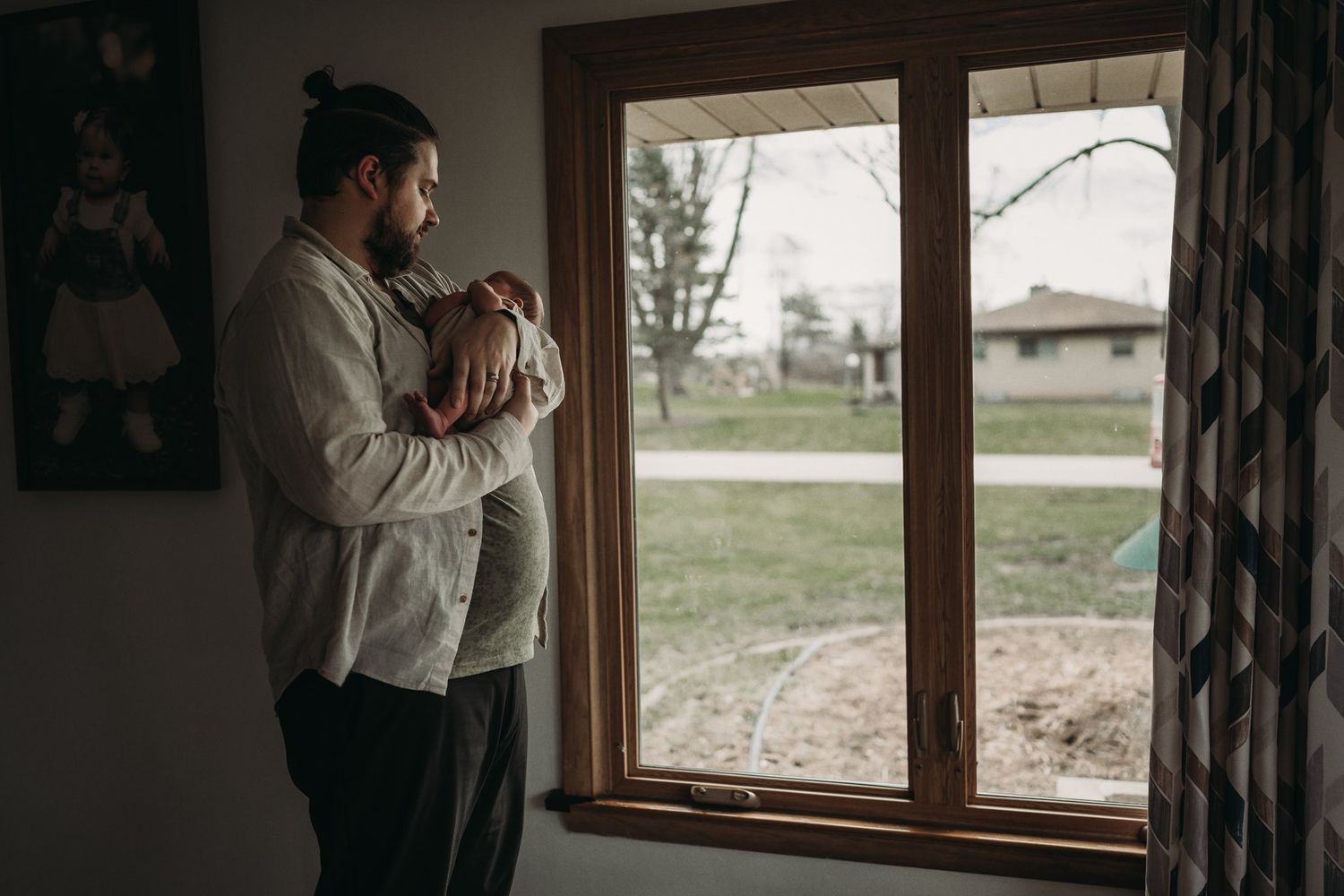 A person stands by a window holding a baby while looking out at the view beyond.