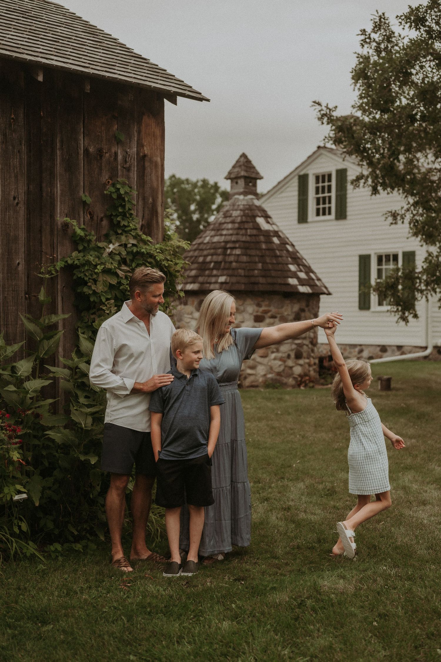 Family plays together near rustic barn and white farmhouse on cloudy summer day.