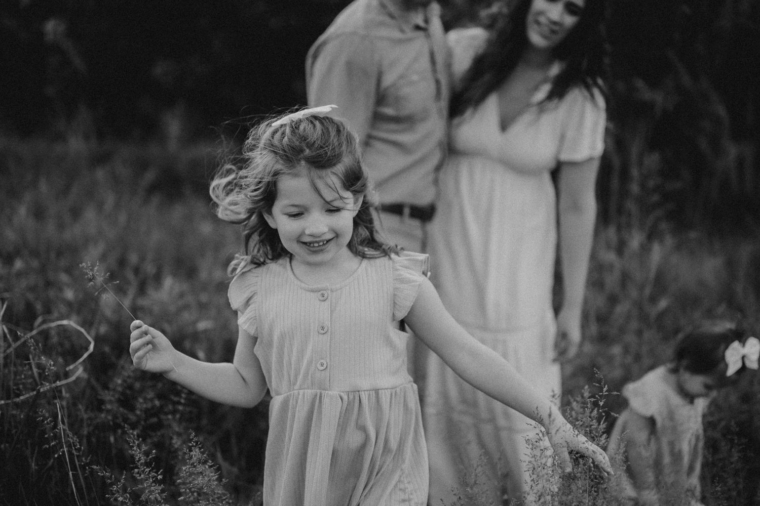 Black and white capture of joyful child dancing in a meadow while others watch in background.