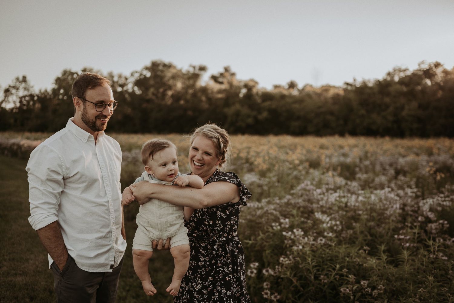 Family stands together in wildflower field during sunset with treeline in background.