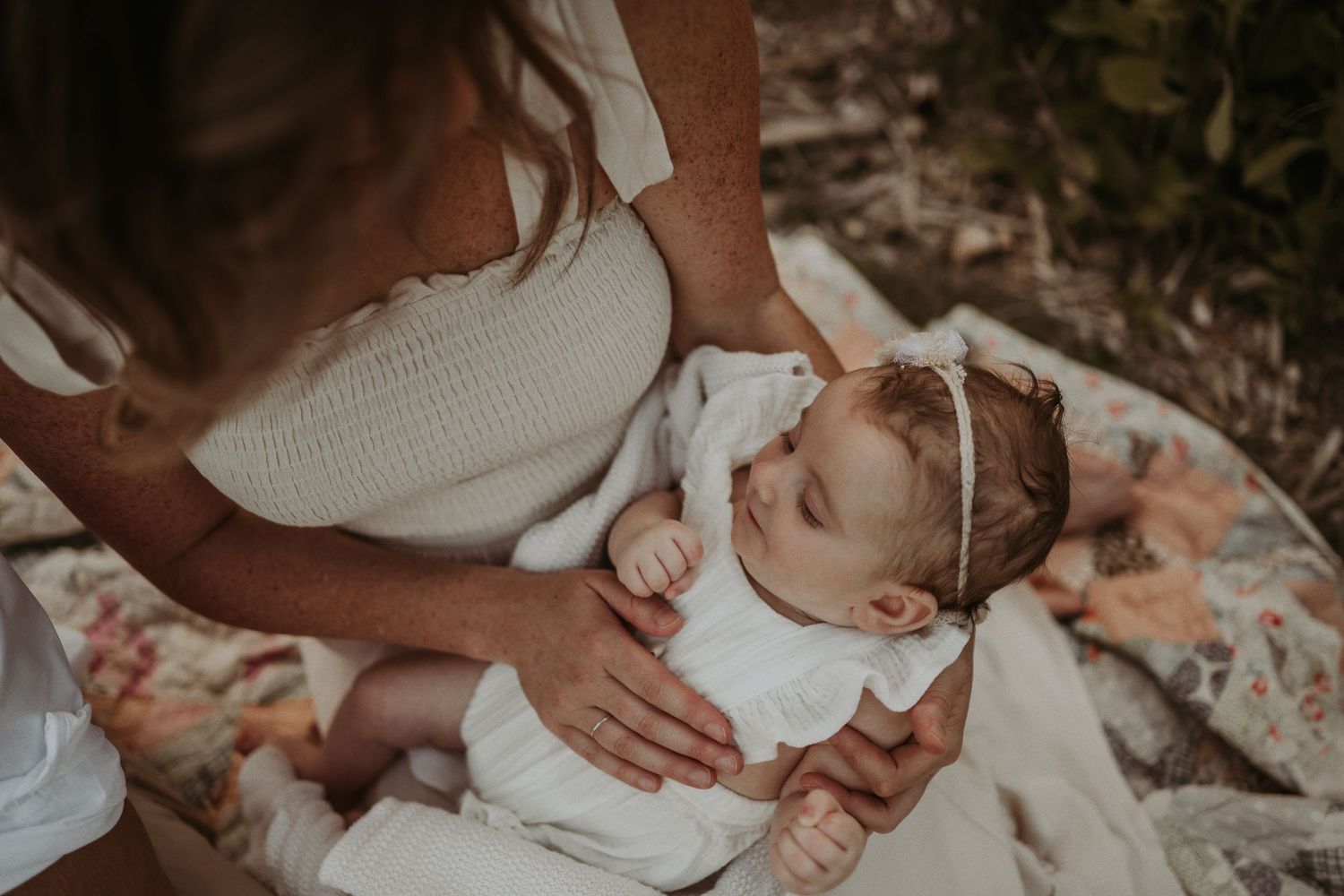 An intimate moment with a baby in white dress and headband lying on a floral blanket.