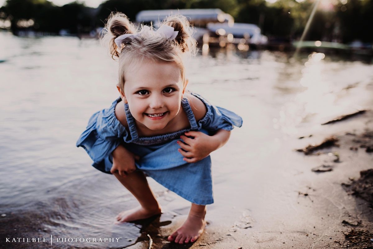 A playful child in a blue dress crouches by the water's edge at sunset.
