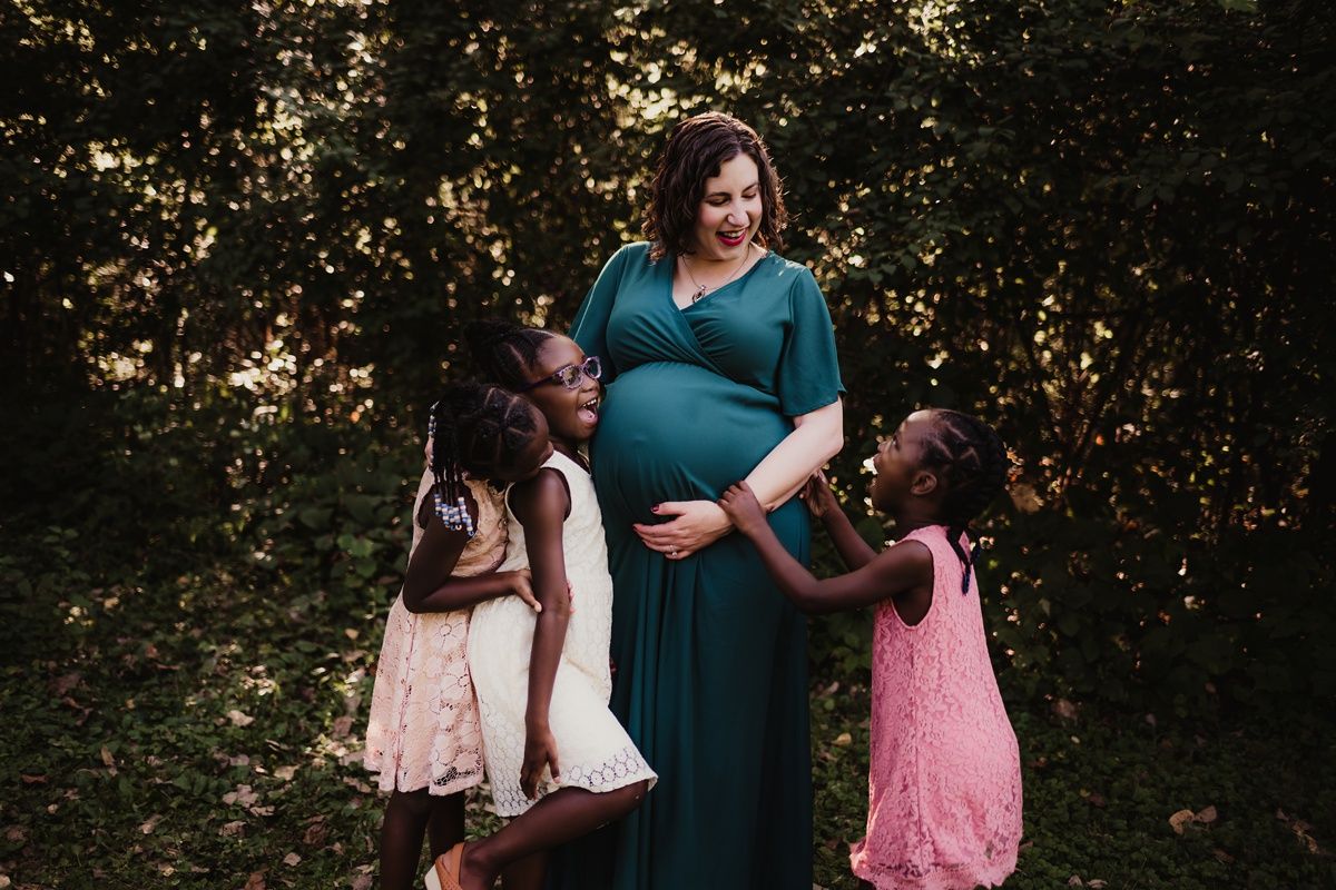Family portrait showing a pregnant woman in teal dress with children in pink and white dresses outdoors.
