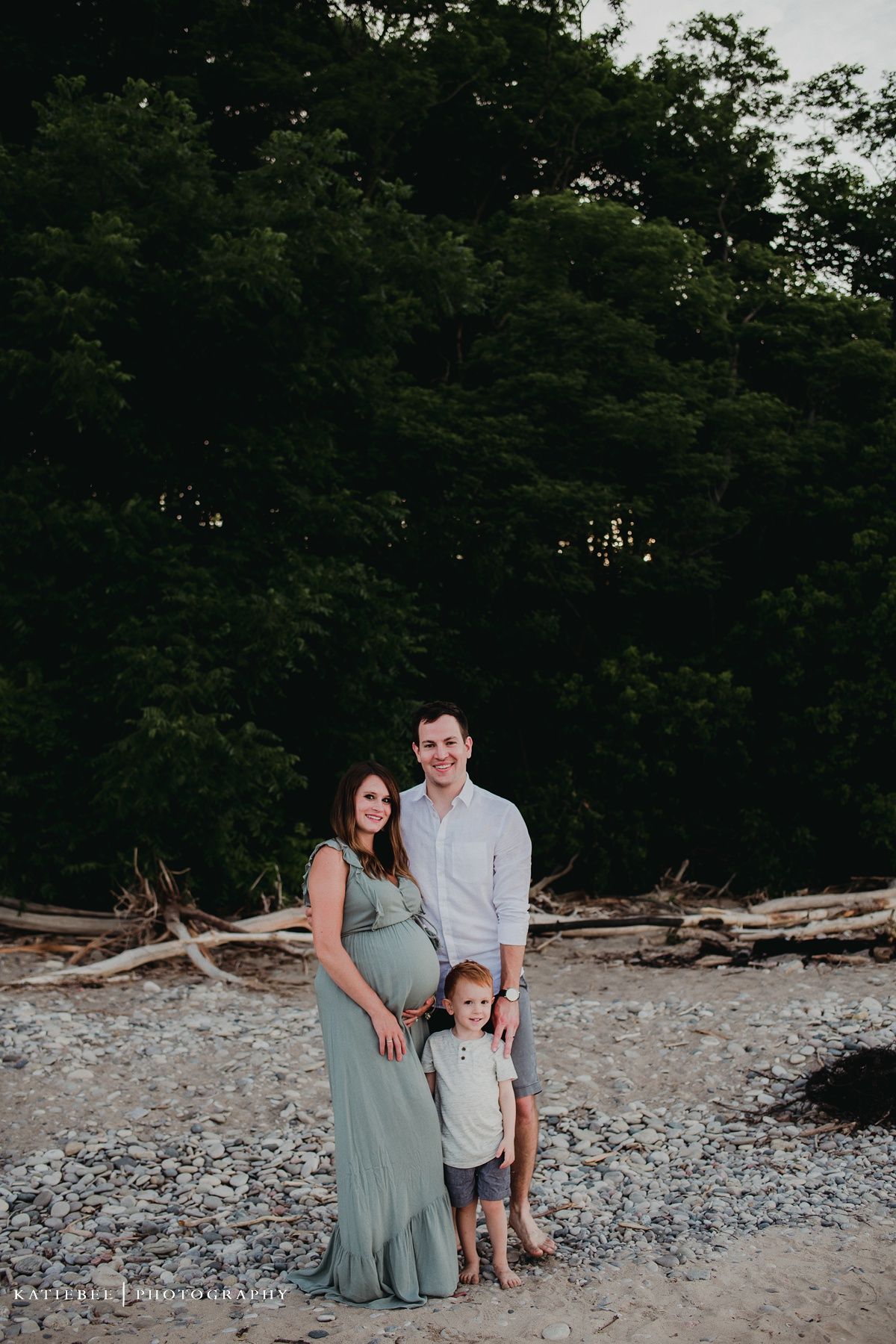 Family portrait on a pebble beach with towering trees creating a natural backdrop.