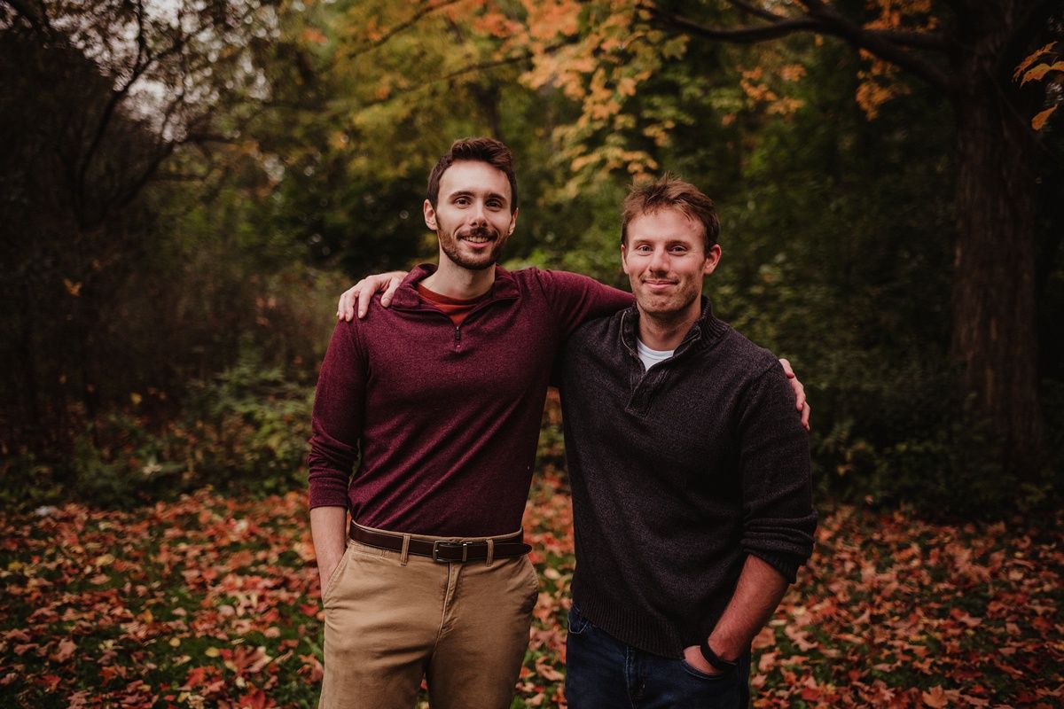 Two people in casual wear standing together against autumn forest backdrop.