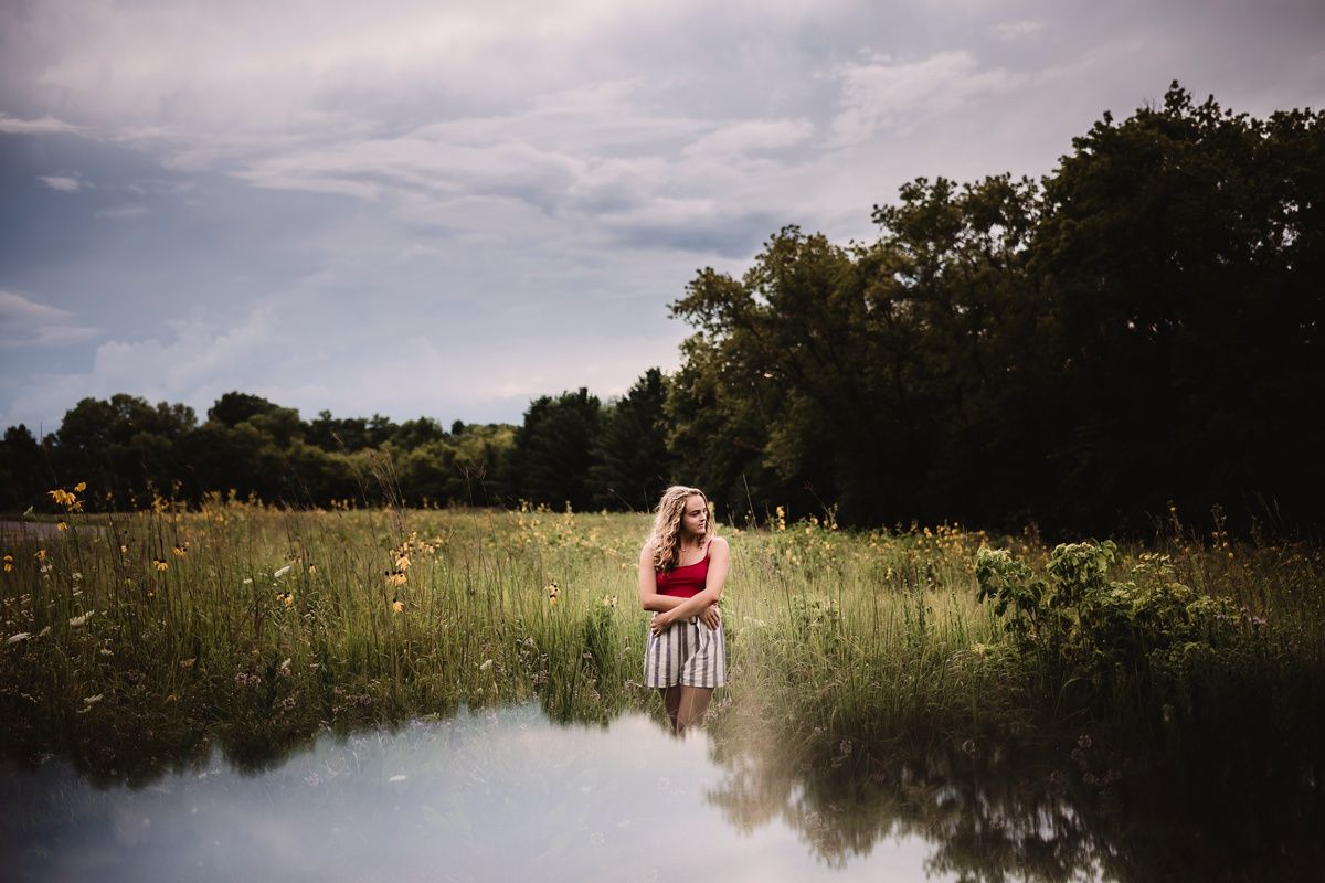 Reflective portrait in a summer meadow with trees and cloudy sky mirrored in water.