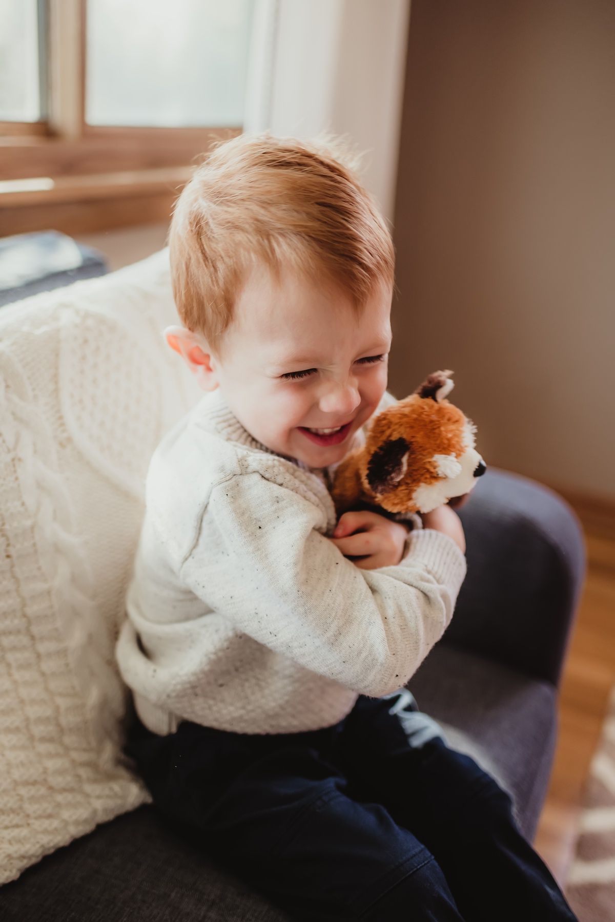 Happy toddler in white sweater laughing while holding stuffed fox toy.