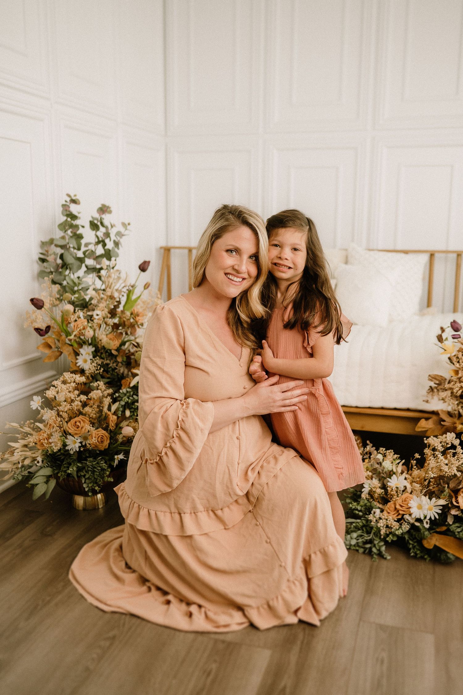 Two people in matching peach-colored dresses pose together in a room decorated with floral arrangements and white walls.