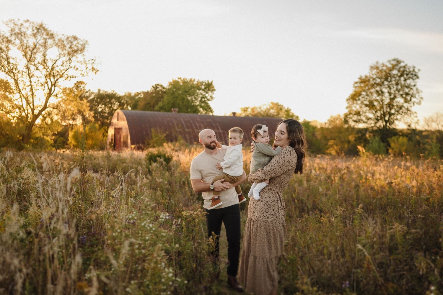 A family stands together in a golden field at sunset during autumn with trees in the background.