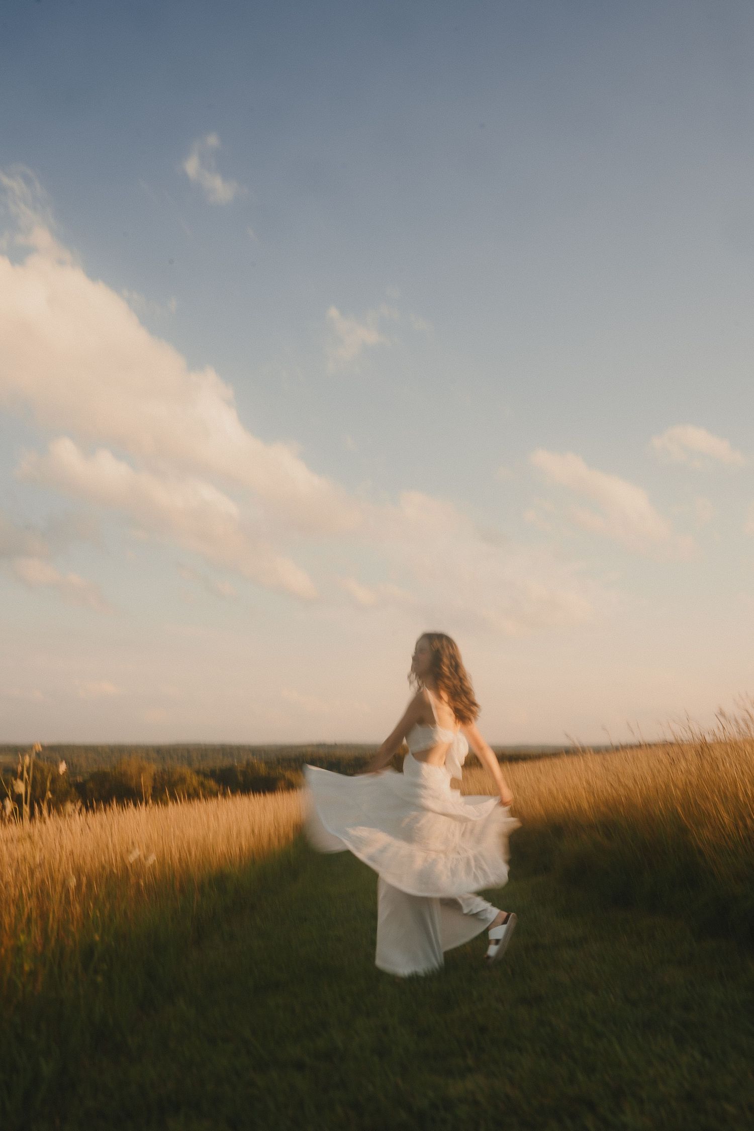 A person in a flowing white dress twirls on a grassy hilltop during golden hour with soft clouds in the sky.