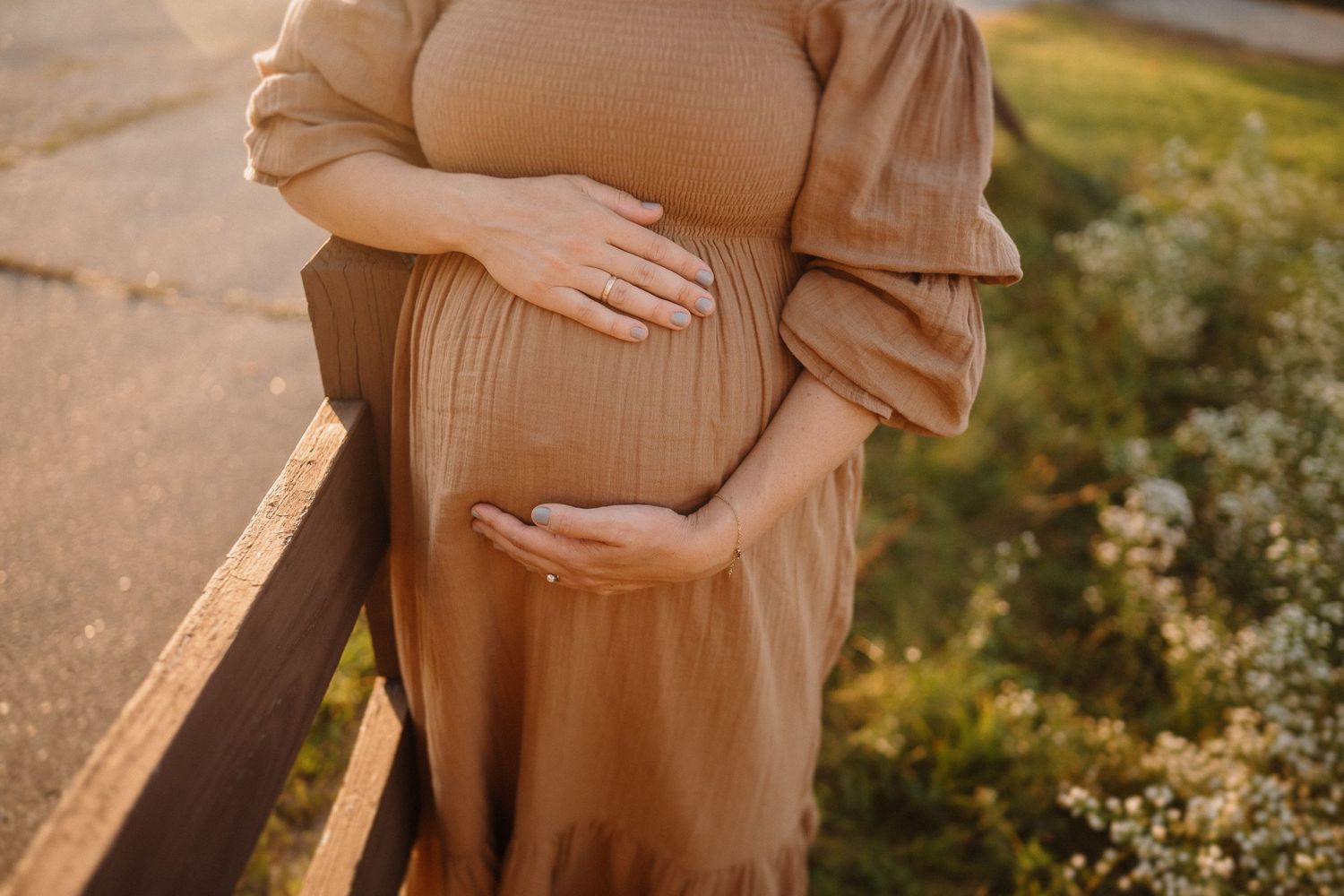 Close-up of hands cradling pregnant belly in brown dress at sunset.