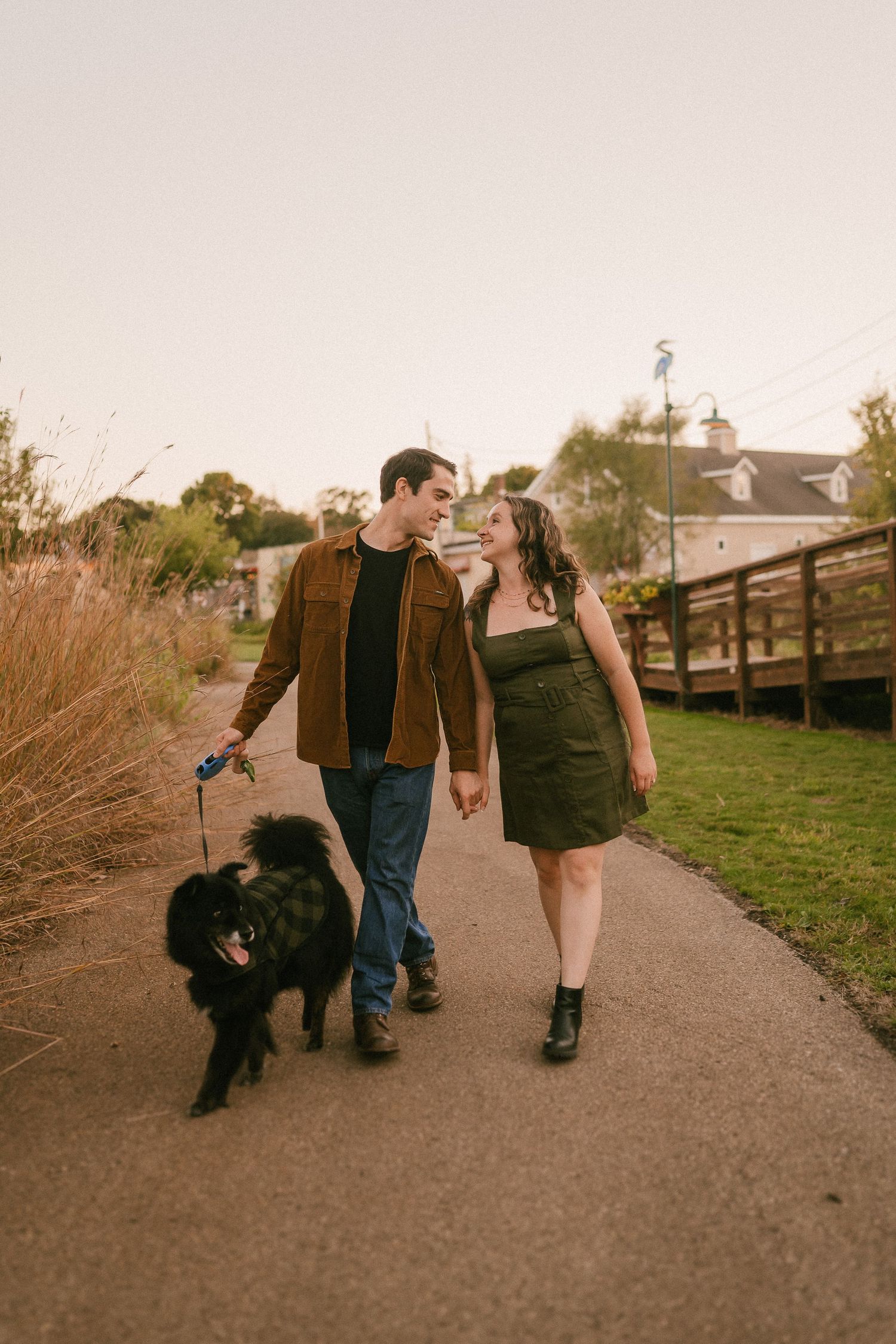 A couple strolls with their black dog on a paved path during golden hour.