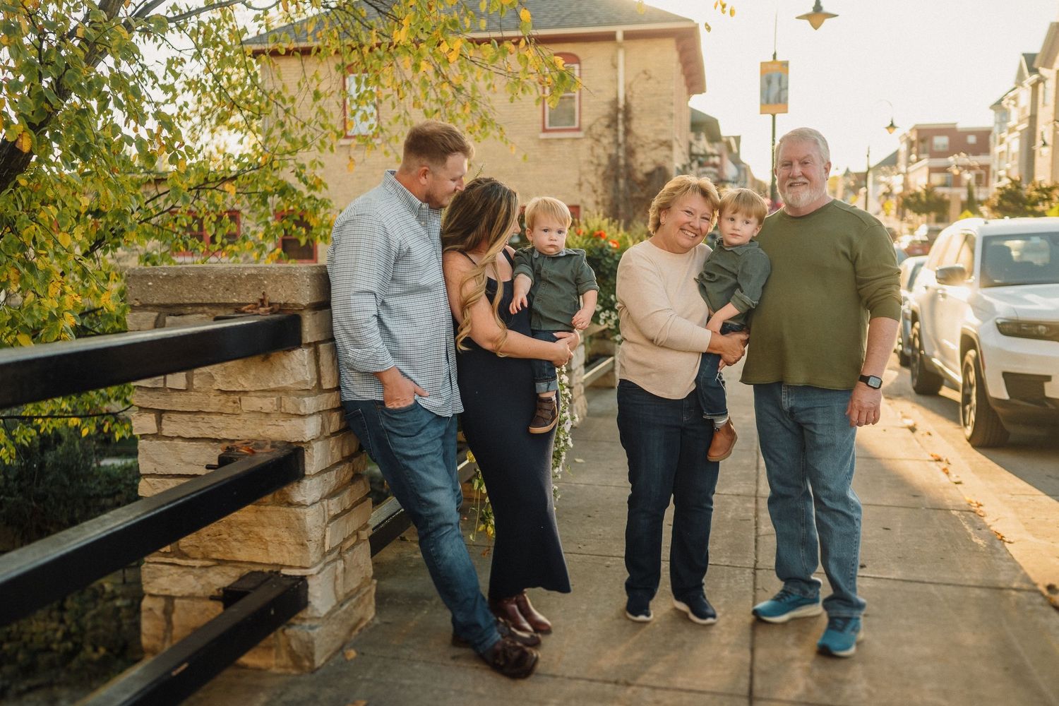 Multi-generational family gathering outdoors on an urban sidewalk beside stone wall during golden hour.
