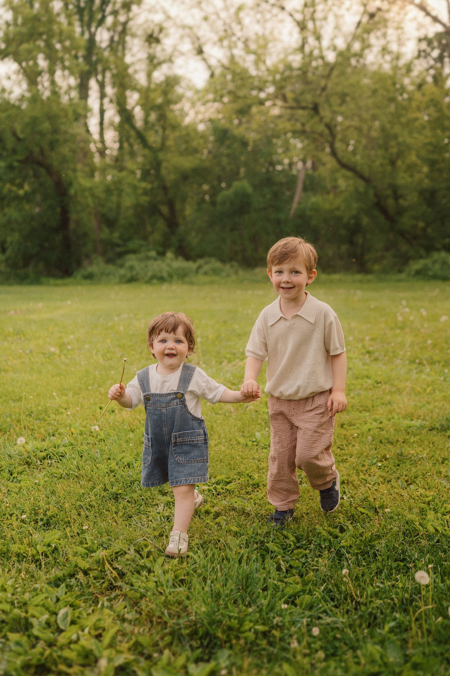 Two young siblings hold hands while playing together in a grassy field with trees in the background.