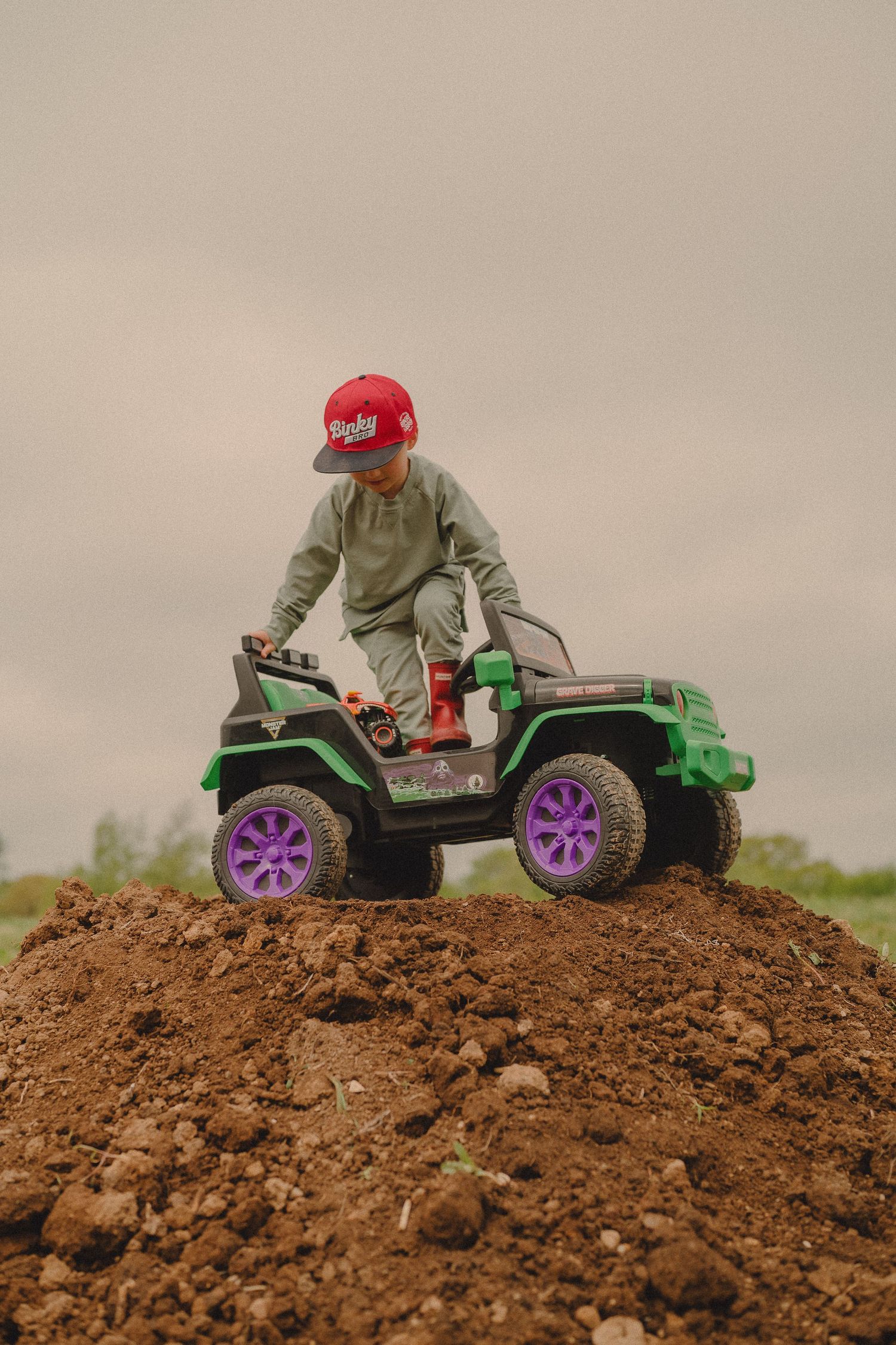 Young boy in red cap riding green ride-on jeep toy over dirt mound on cloudy day.