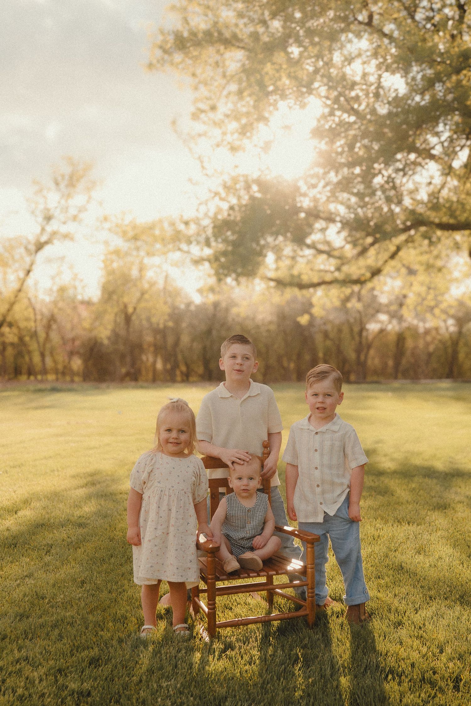 Four children pose outdoors in golden hour light, three boys and a girl in coordinating outfits on a lush green lawn.