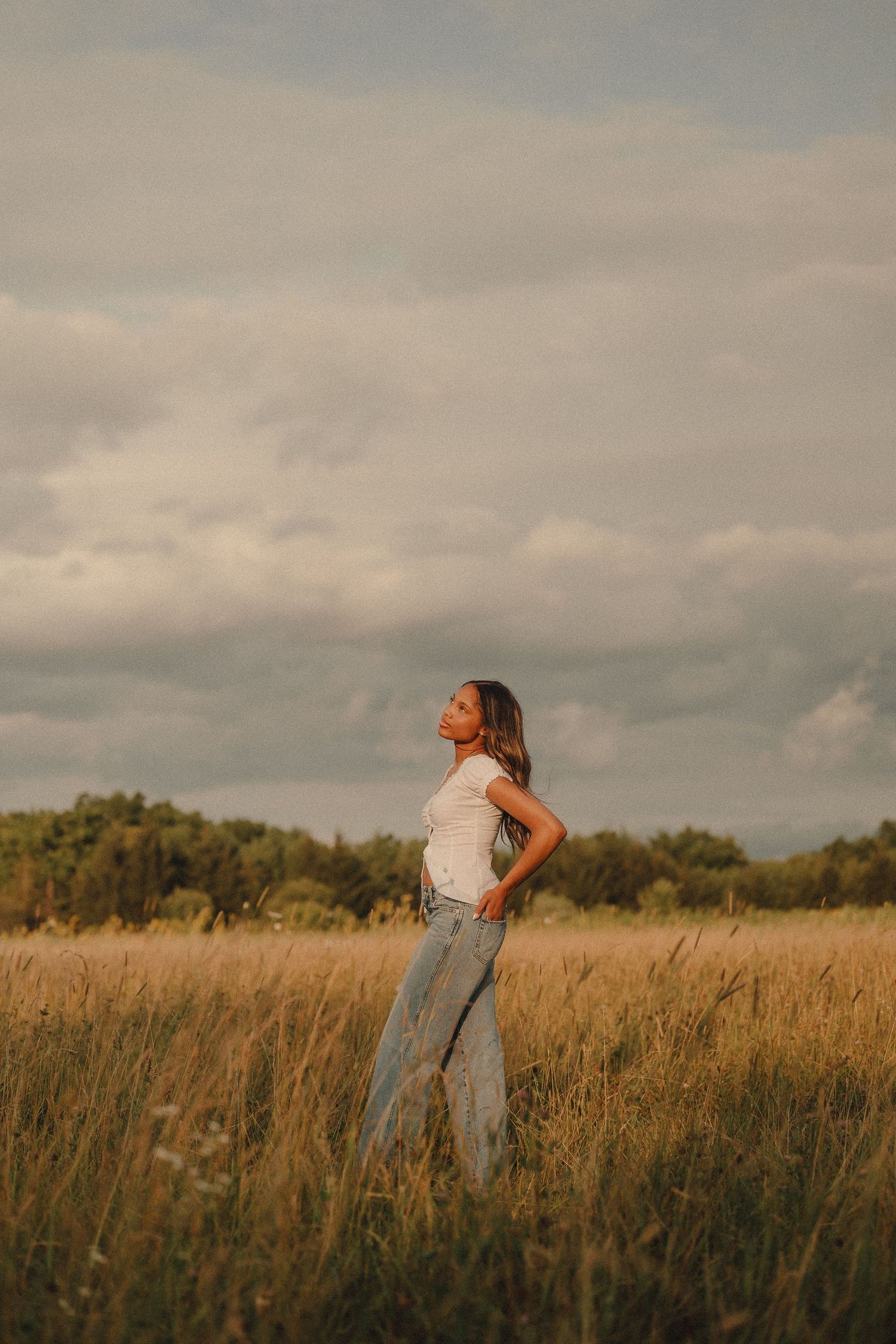 Young woman in white top and jeans standing in golden field under dramatic cloudy sky.