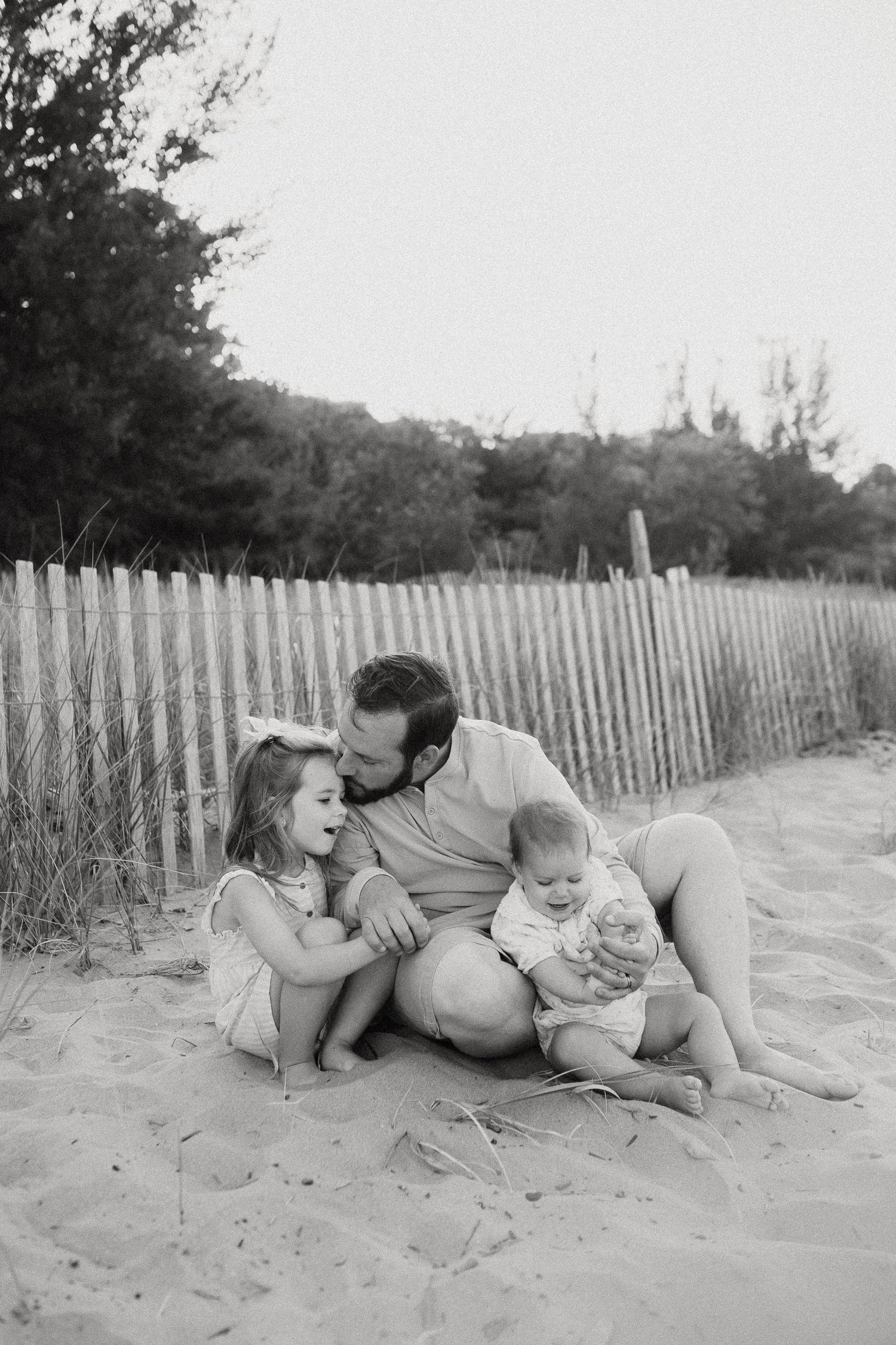 Black and white photo of father kissing daughter while holding baby on beach.