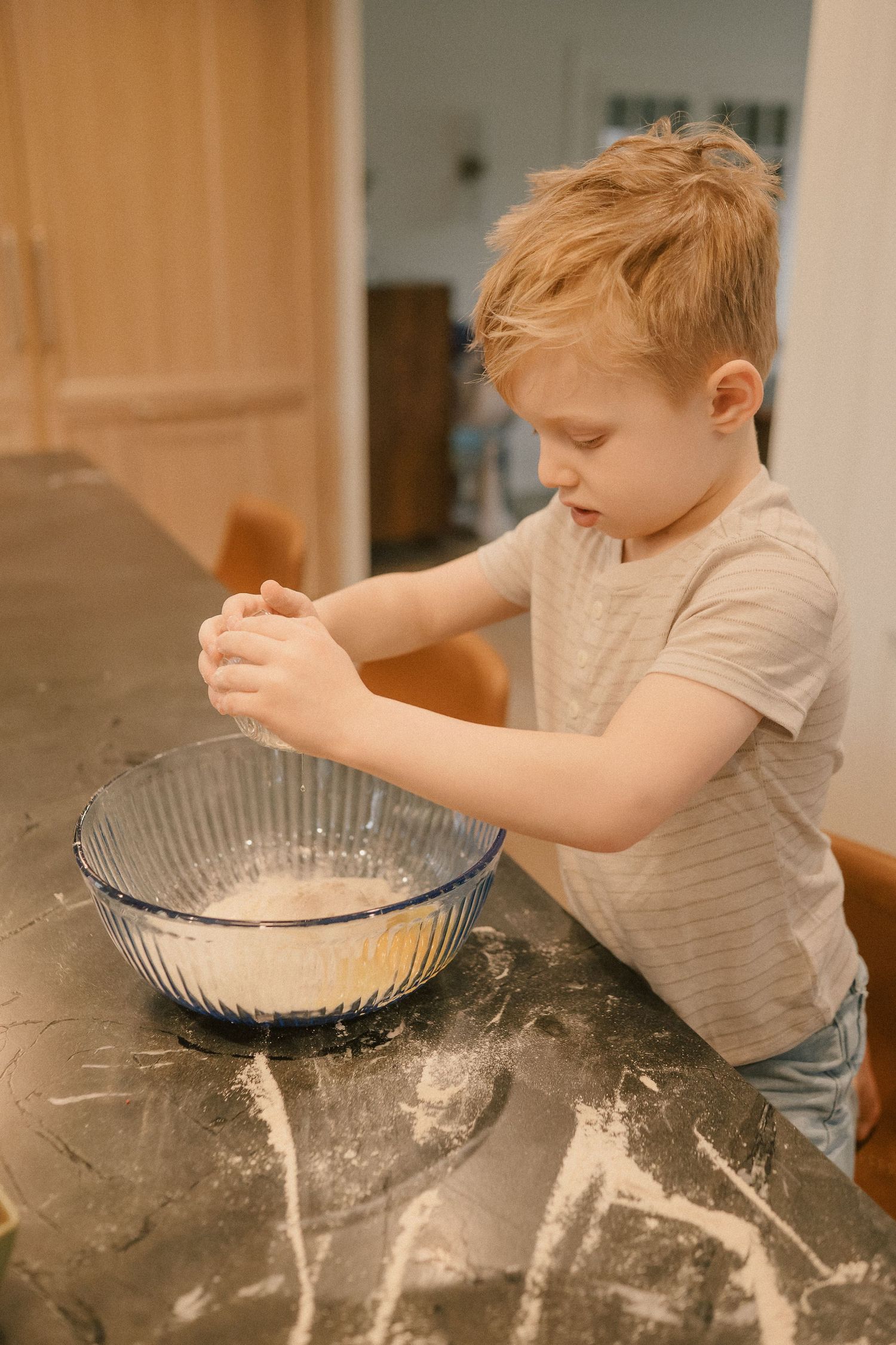 Young boy mixing dough in a glass bowl on a flour-dusted kitchen counter.