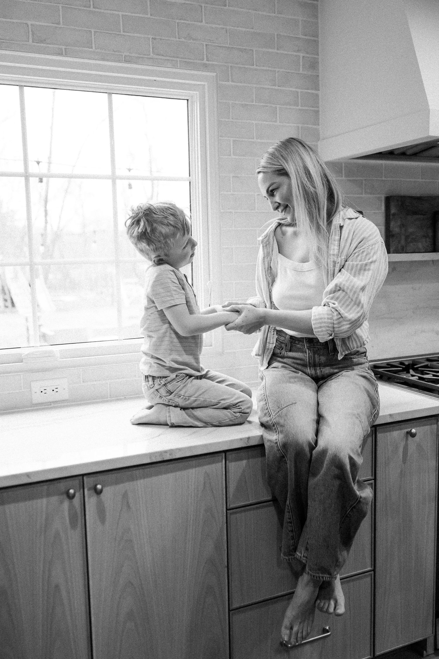 Black and white photo of a mother and young son sitting on a kitchen counter by a window.