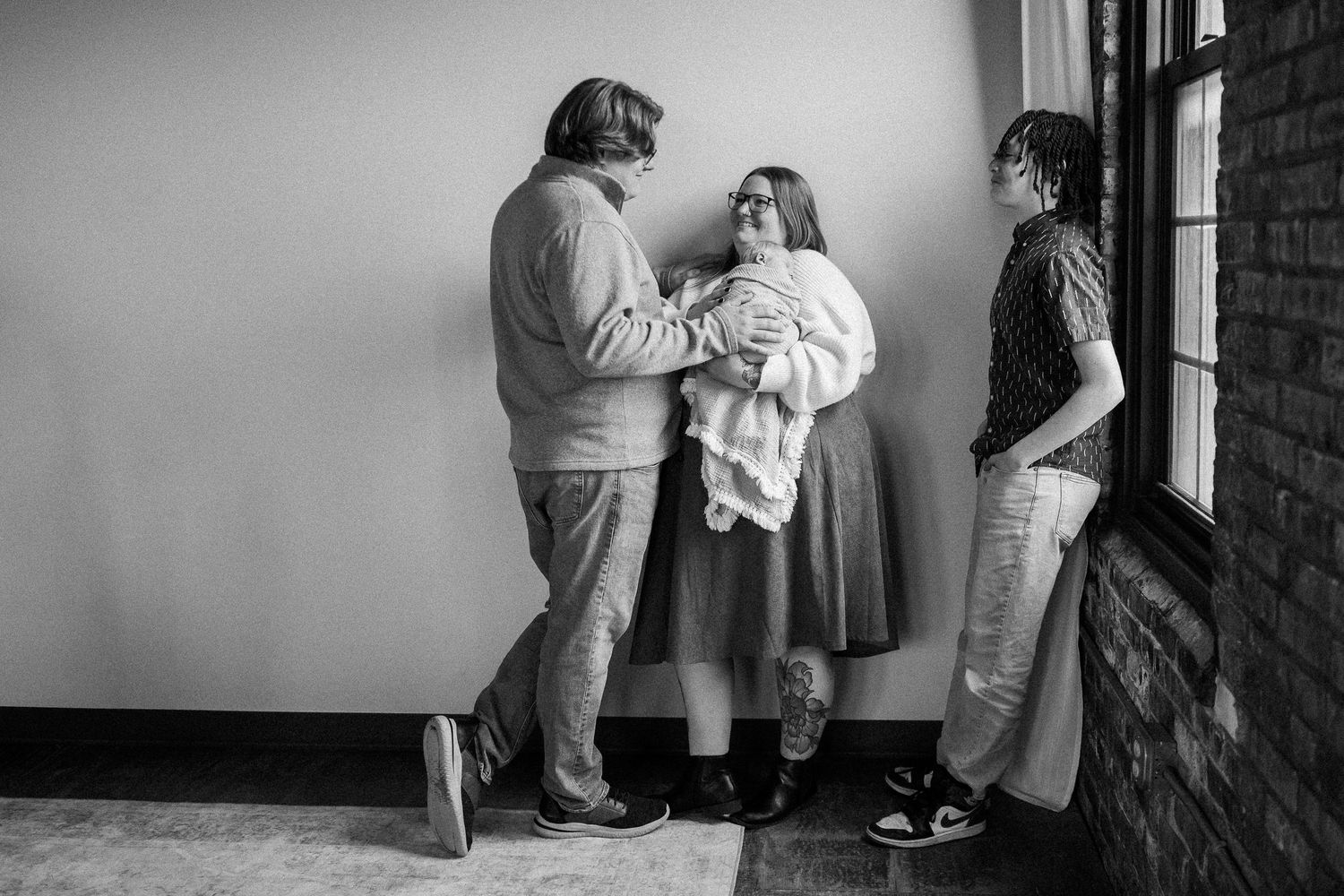 Black and white photo of three people indoors, one holding a newborn baby wrapped in a white blanket.