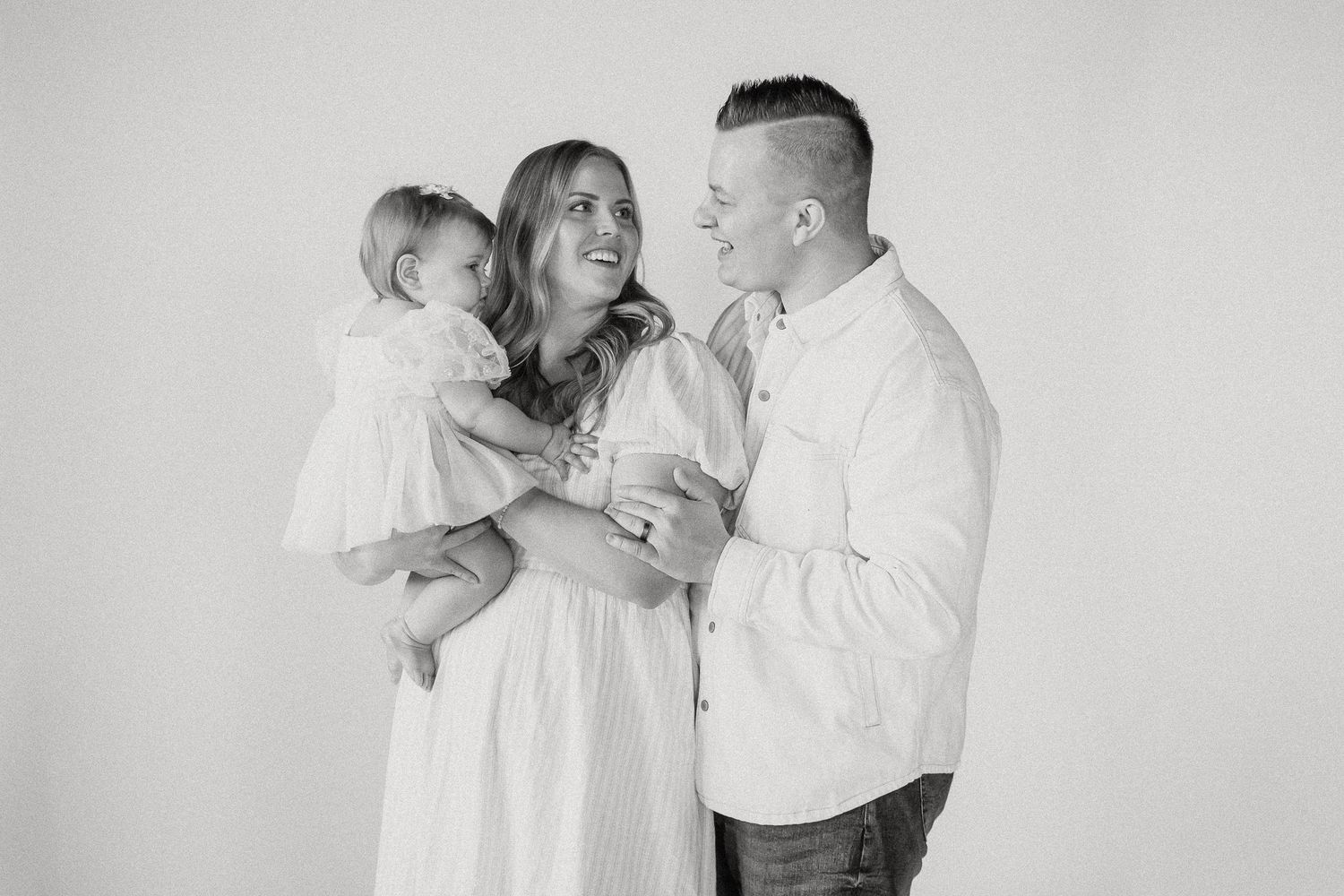 Black and white family portrait of parents smiling at their baby daughter in a bright studio setting.
