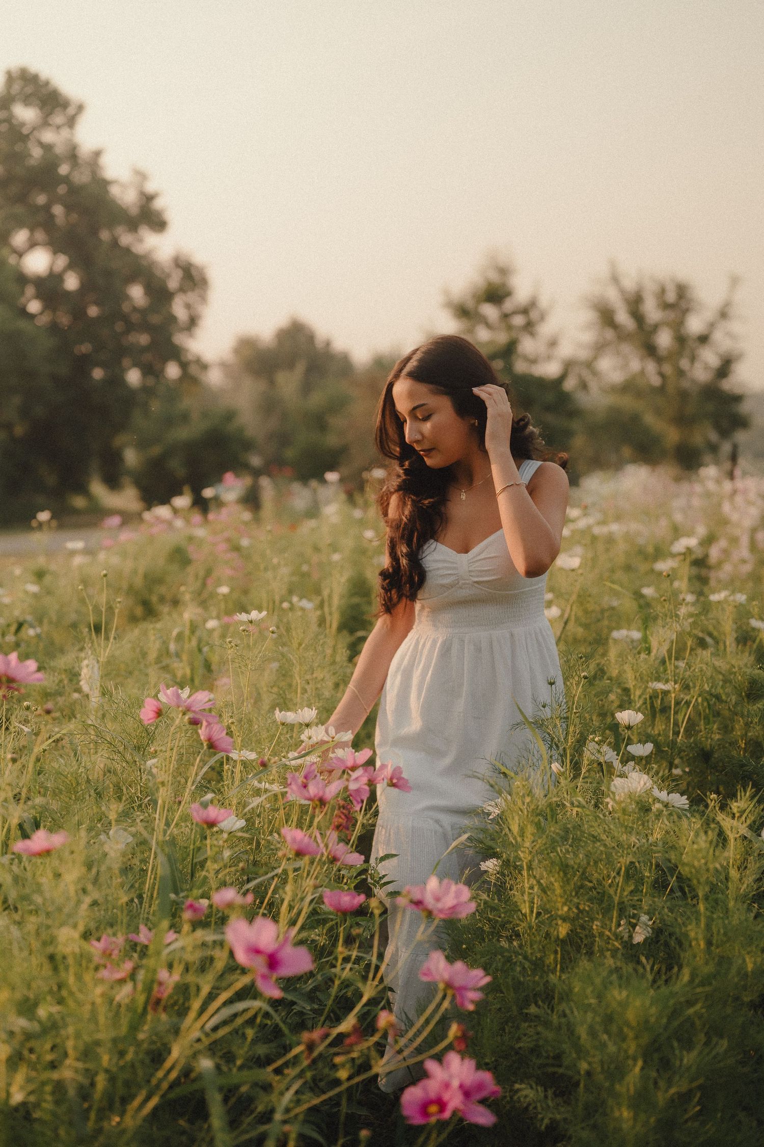 Young woman in white dress walking through pink cosmos flower field at dusk.