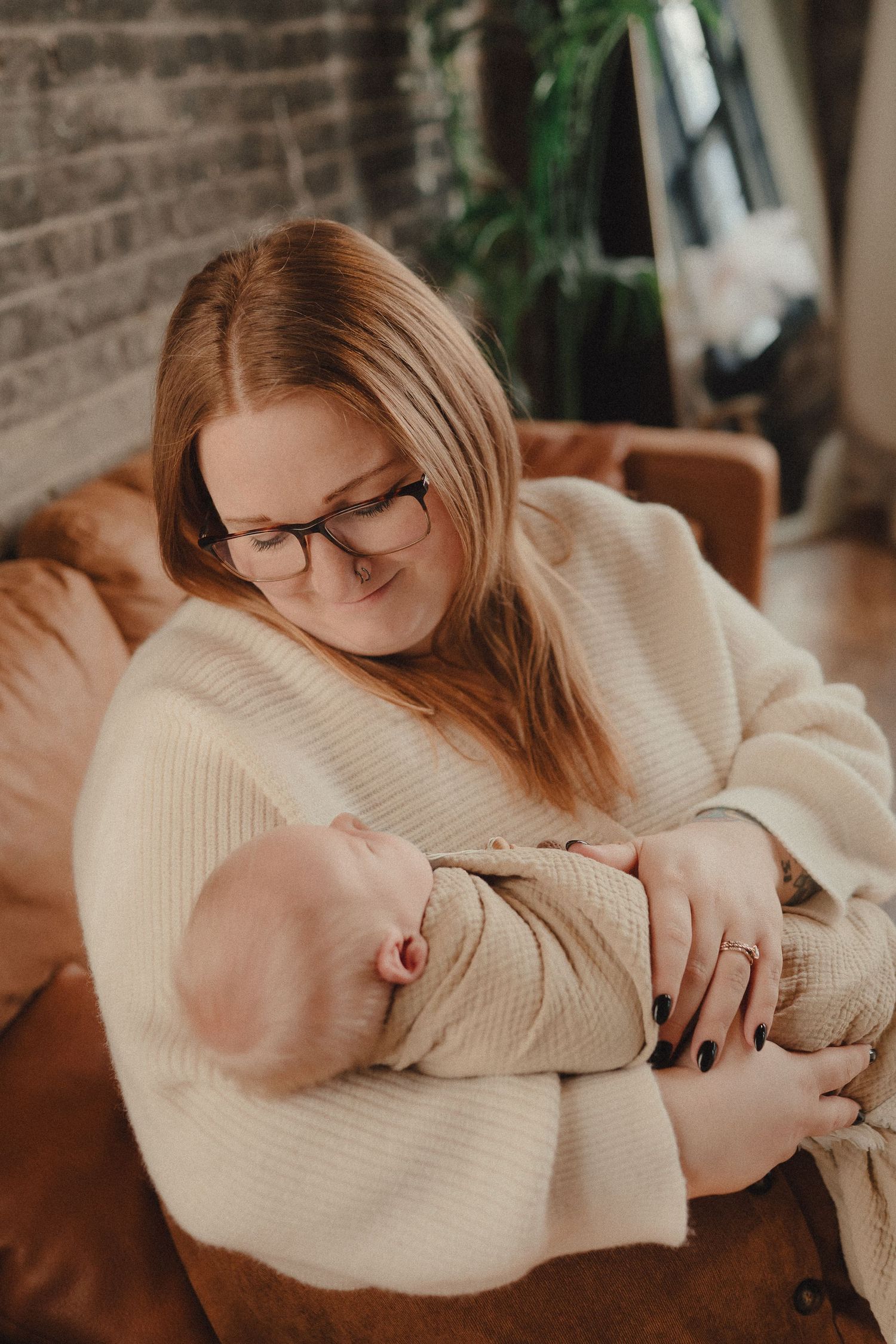 Mother in cream sweater and glasses tenderly holds sleeping newborn baby on brown leather sofa.
