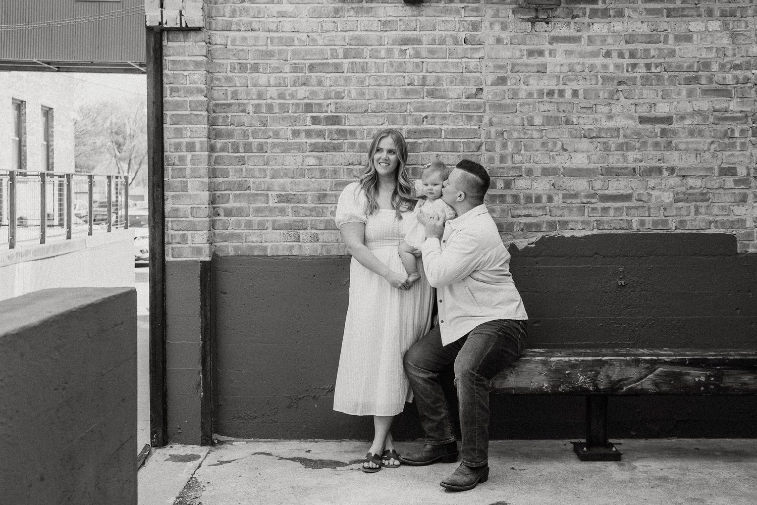 Black and white photo of a couple sharing a tender moment against a brick wall, man kneeling beside pregnant woman in white dress.