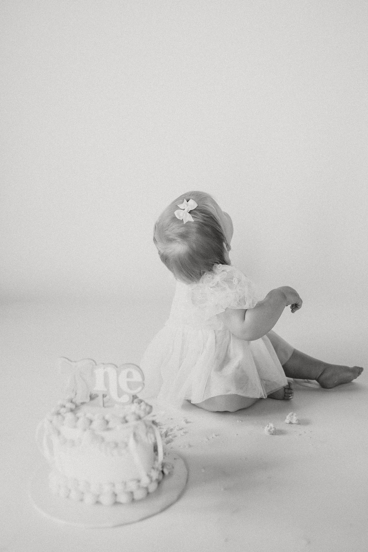 Black and white rear view of a baby girl in a white dress next to her smashed birthday cake.