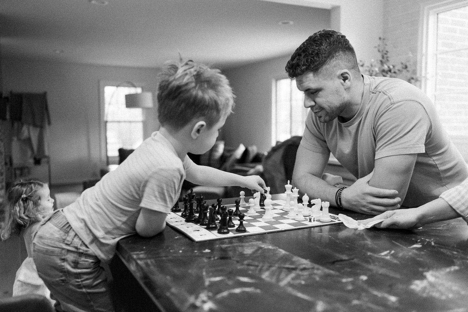 Black and white photo of a father and young son playing chess together at a wooden table.