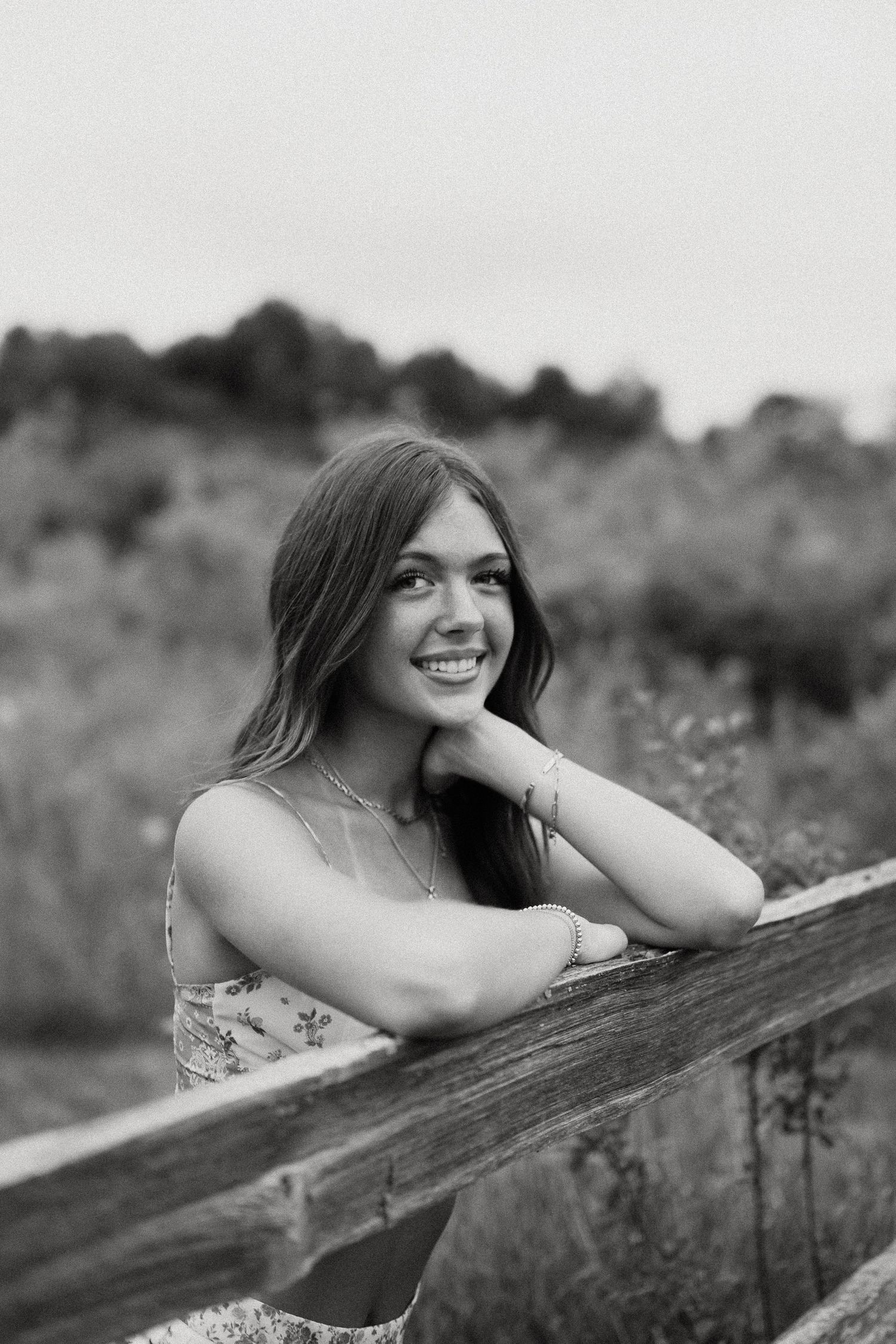 Smiling teenage girl leaning on a wooden fence in a black and white outdoor portrait photo.