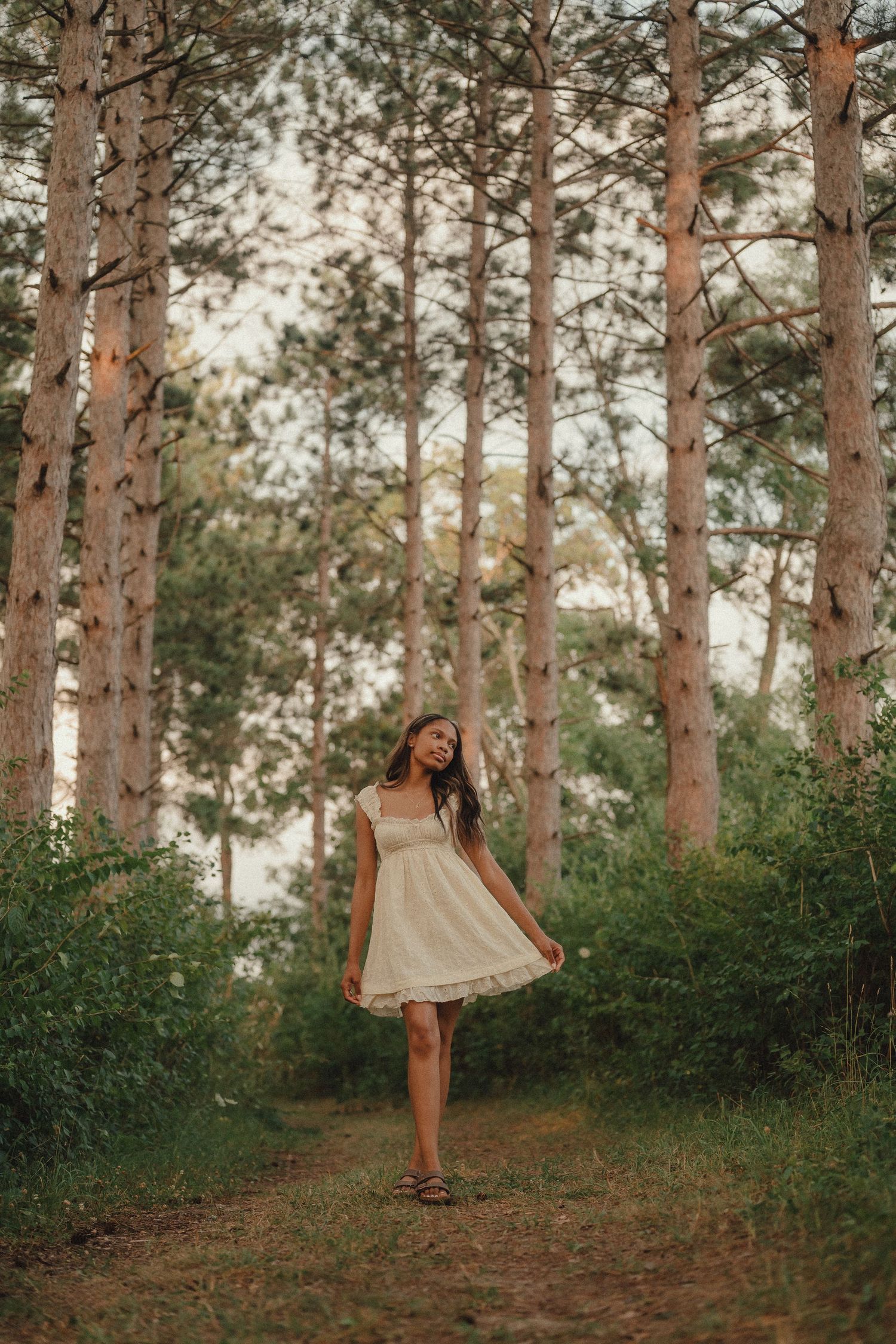 Young woman in white sundress stands on a forest path surrounded by tall pine trees at dusk.