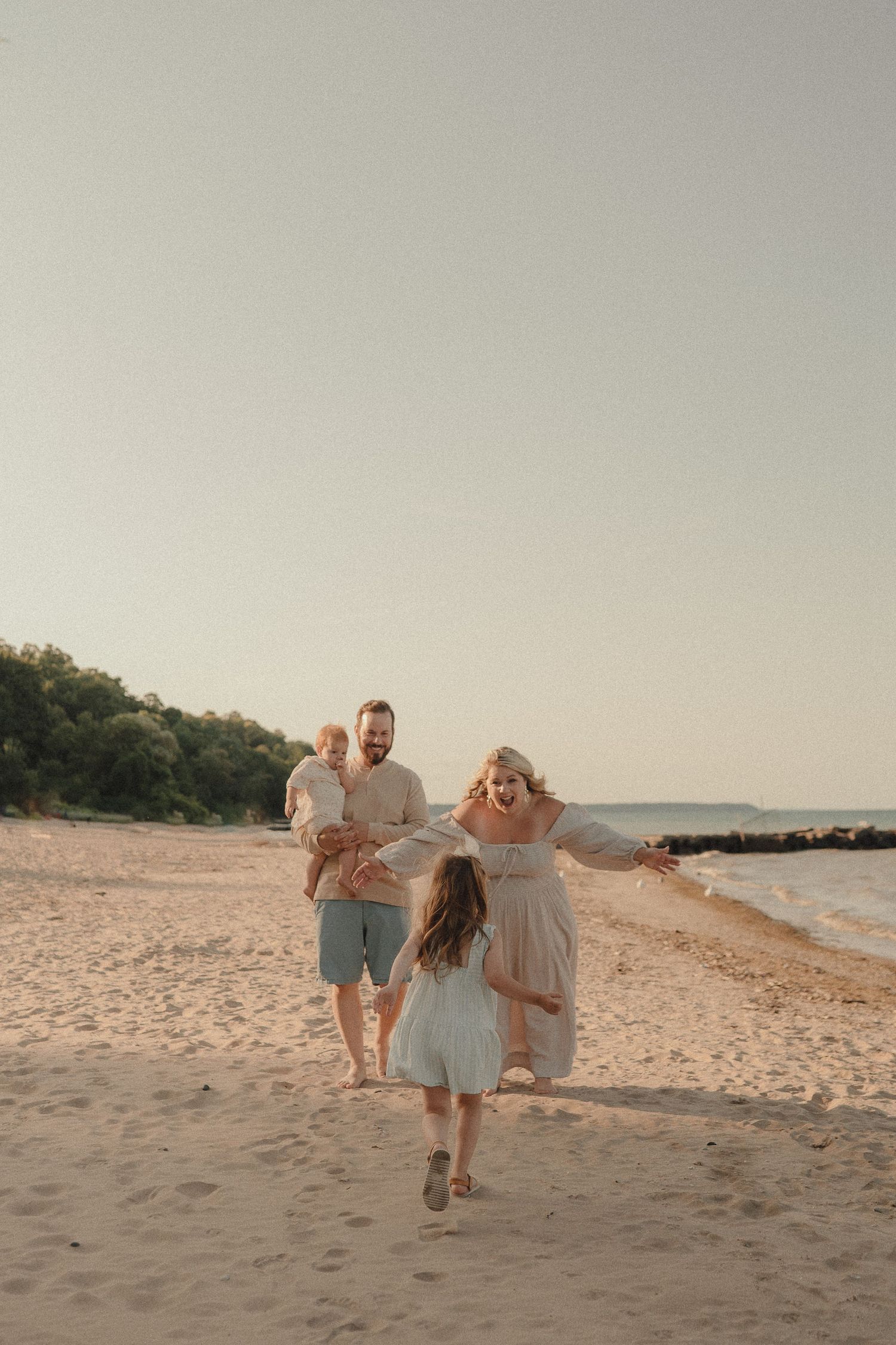 Family of four laughing and playing on a sandy beach at golden hour, mother reaching out to young daughter running toward her.