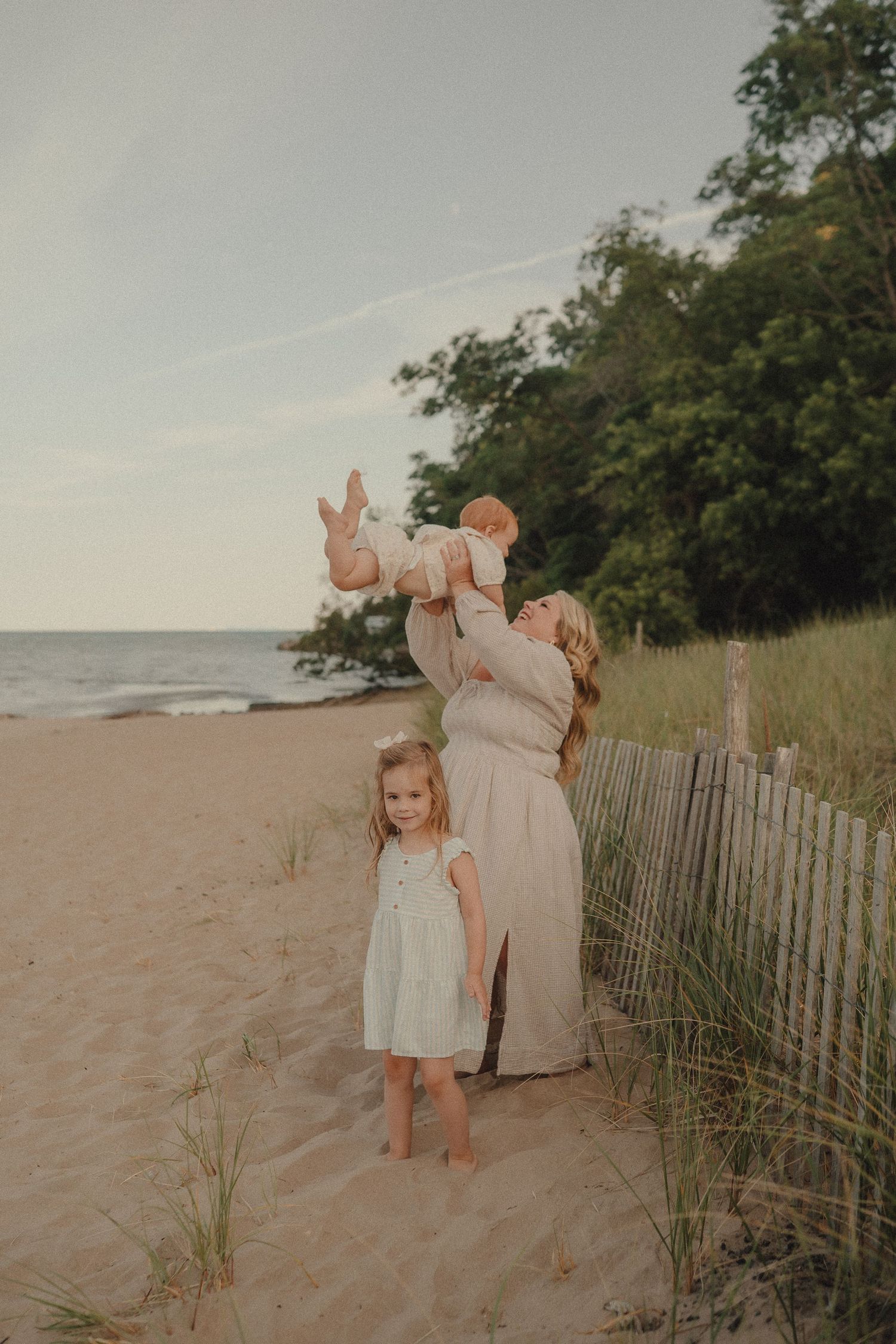 A mother lifts her baby on a sandy beach while a young girl in a white dress stands nearby by a wooden fence.