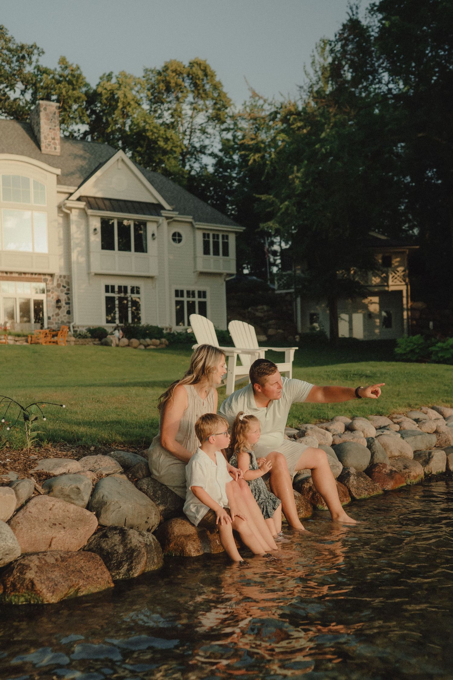 Family of four sitting by a rocky waterfront at golden hour with a large white house in the background.