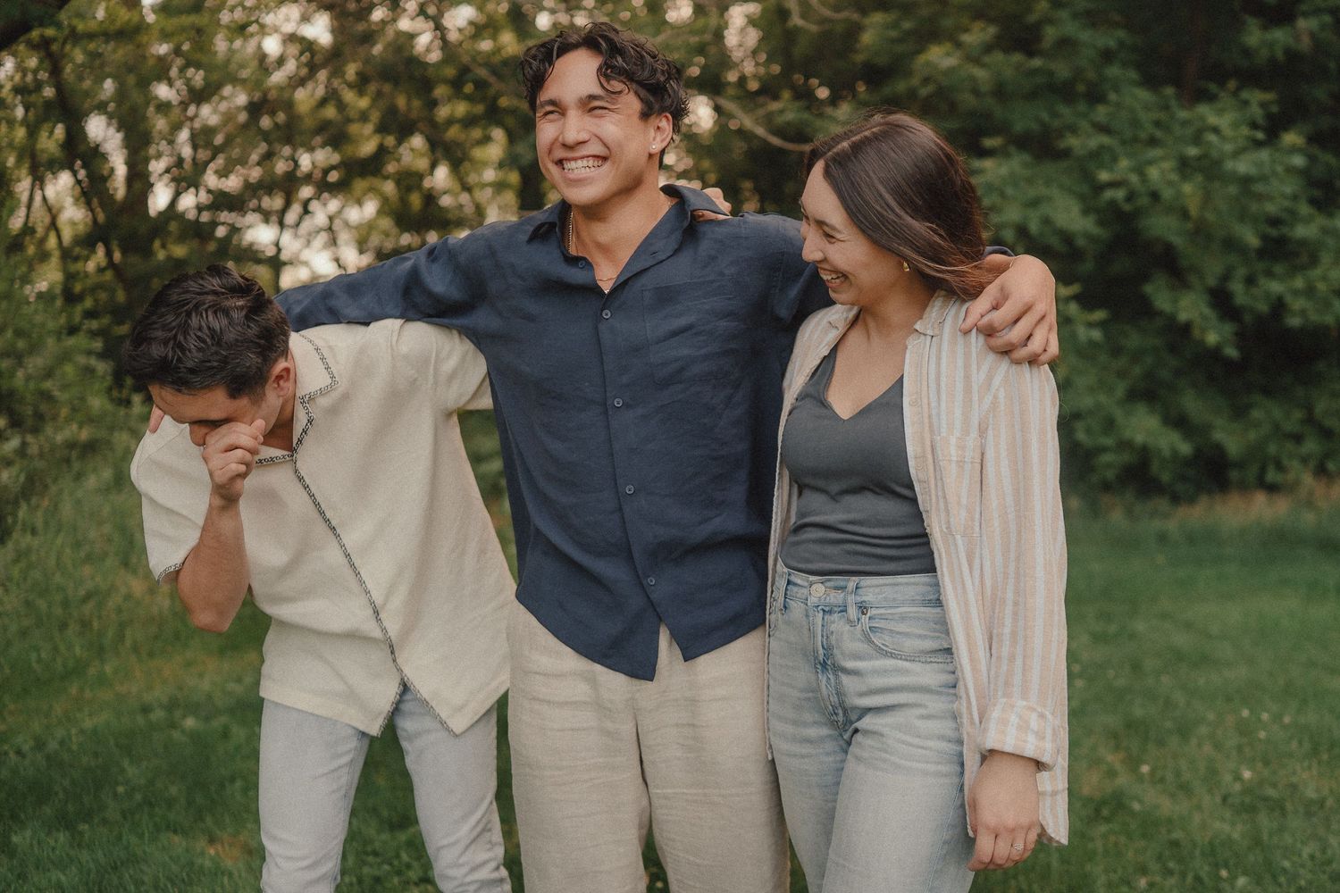 Three friends laughing and embracing outdoors in a lush green park setting, casually dressed in neutral tones.