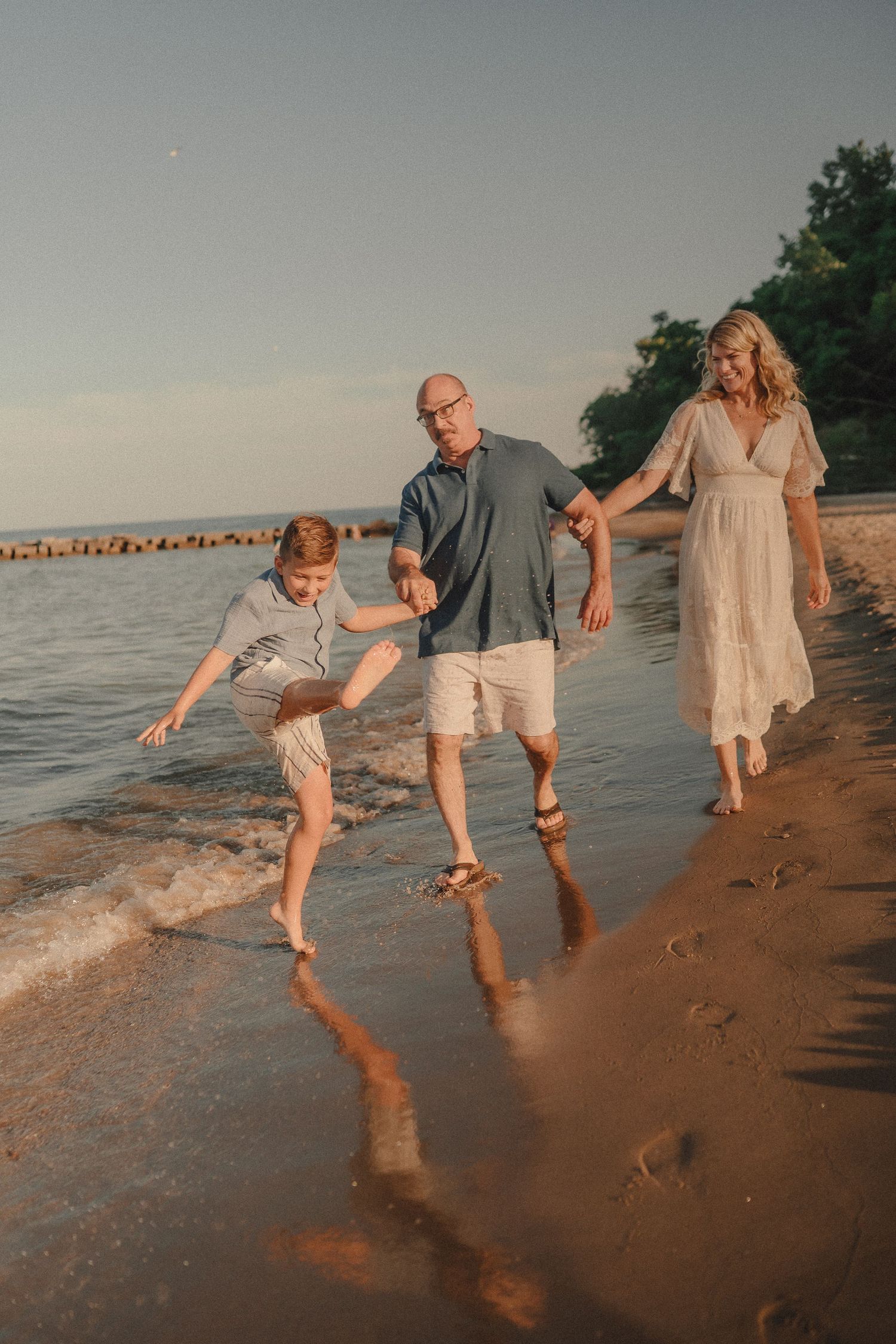 Family of three walking along beach shoreline at golden hour, boy splashing in waves.