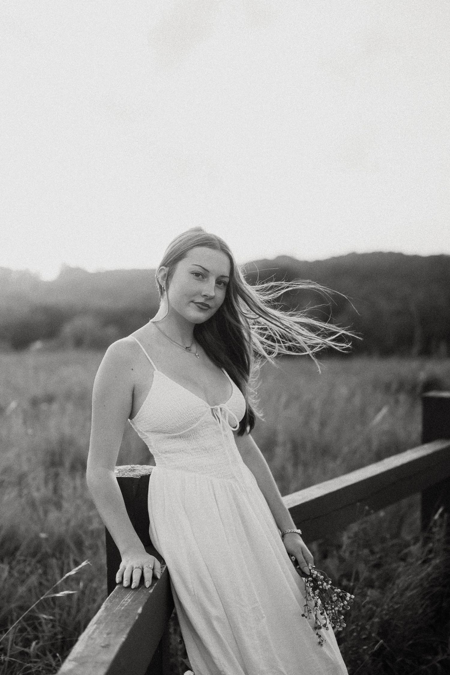Young woman in white sundress leaning on wooden fence in field, hair blowing in wind, black and white photo.