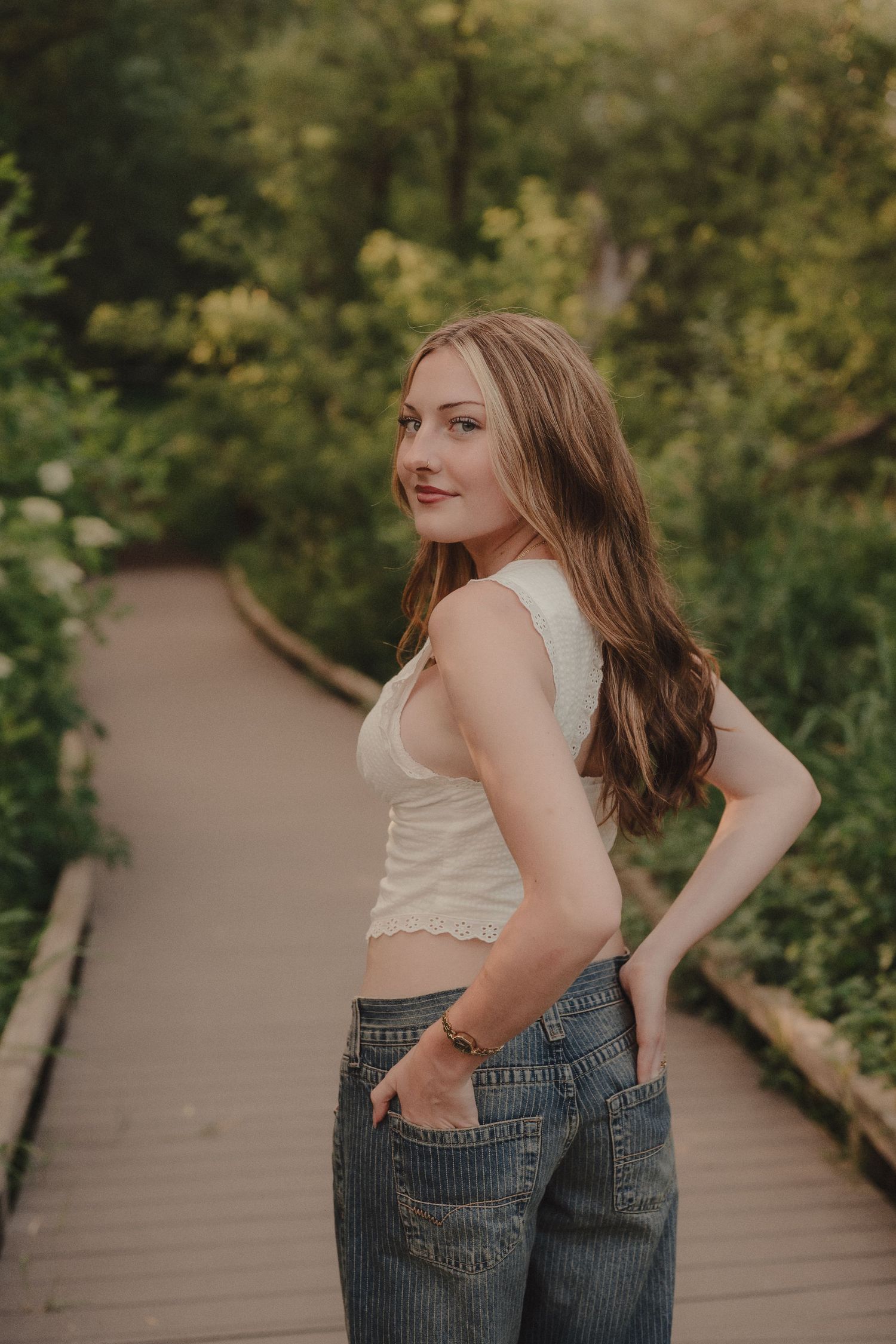 Young woman in white tank top and wide-leg jeans stands on a garden boardwalk path.