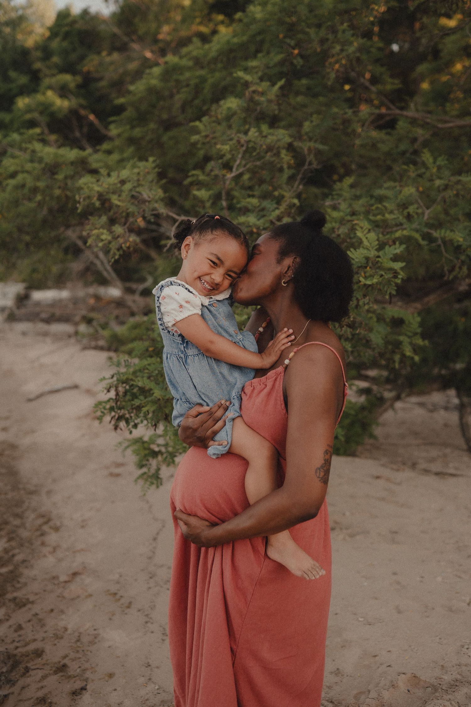 Glowing pregnant mother in coral dress kissing smiling toddler daughter on sandy beach near trees.