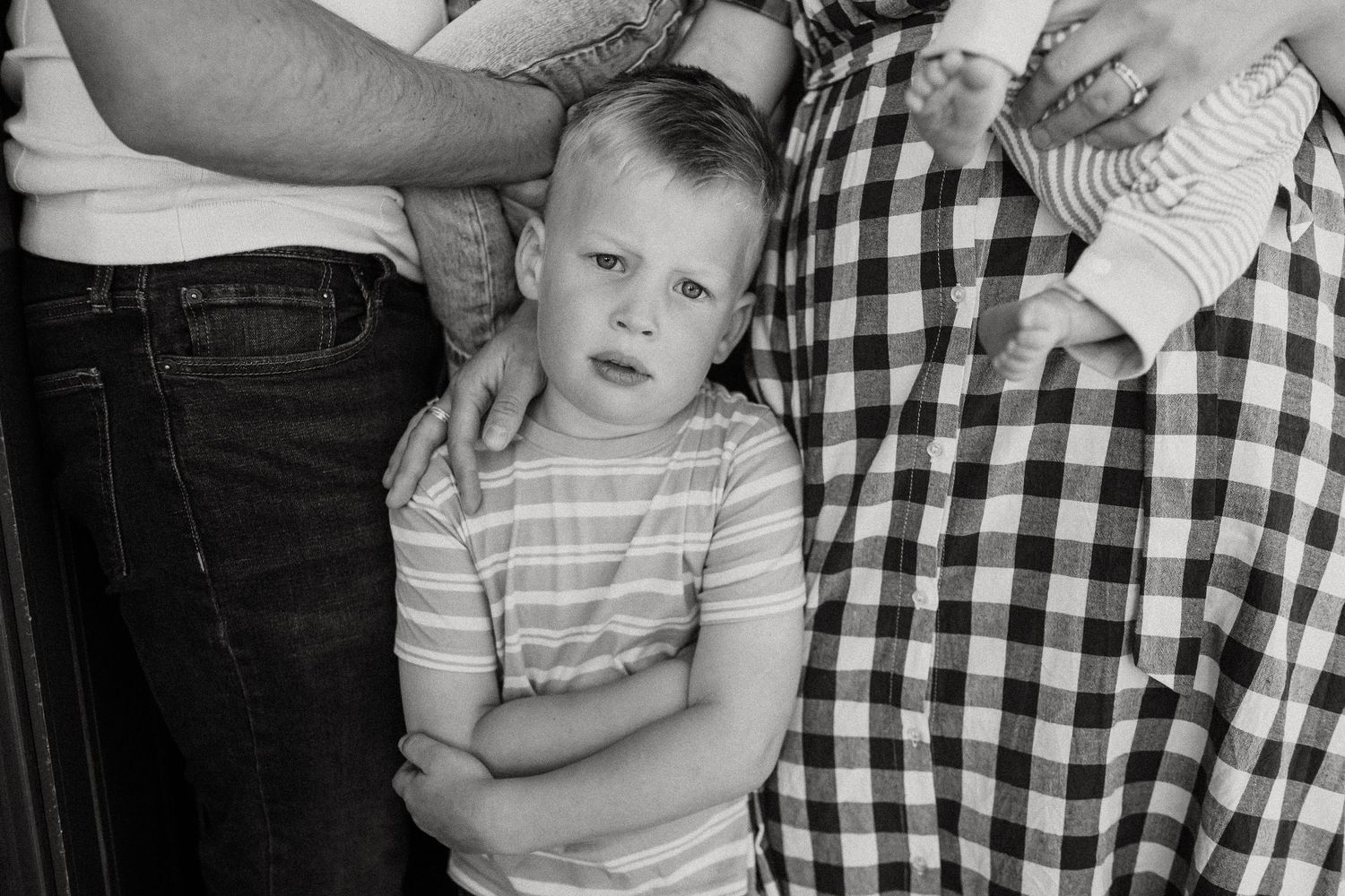 A young boy with crossed arms stands between his parents next to a newborn baby in a black and white portrait.