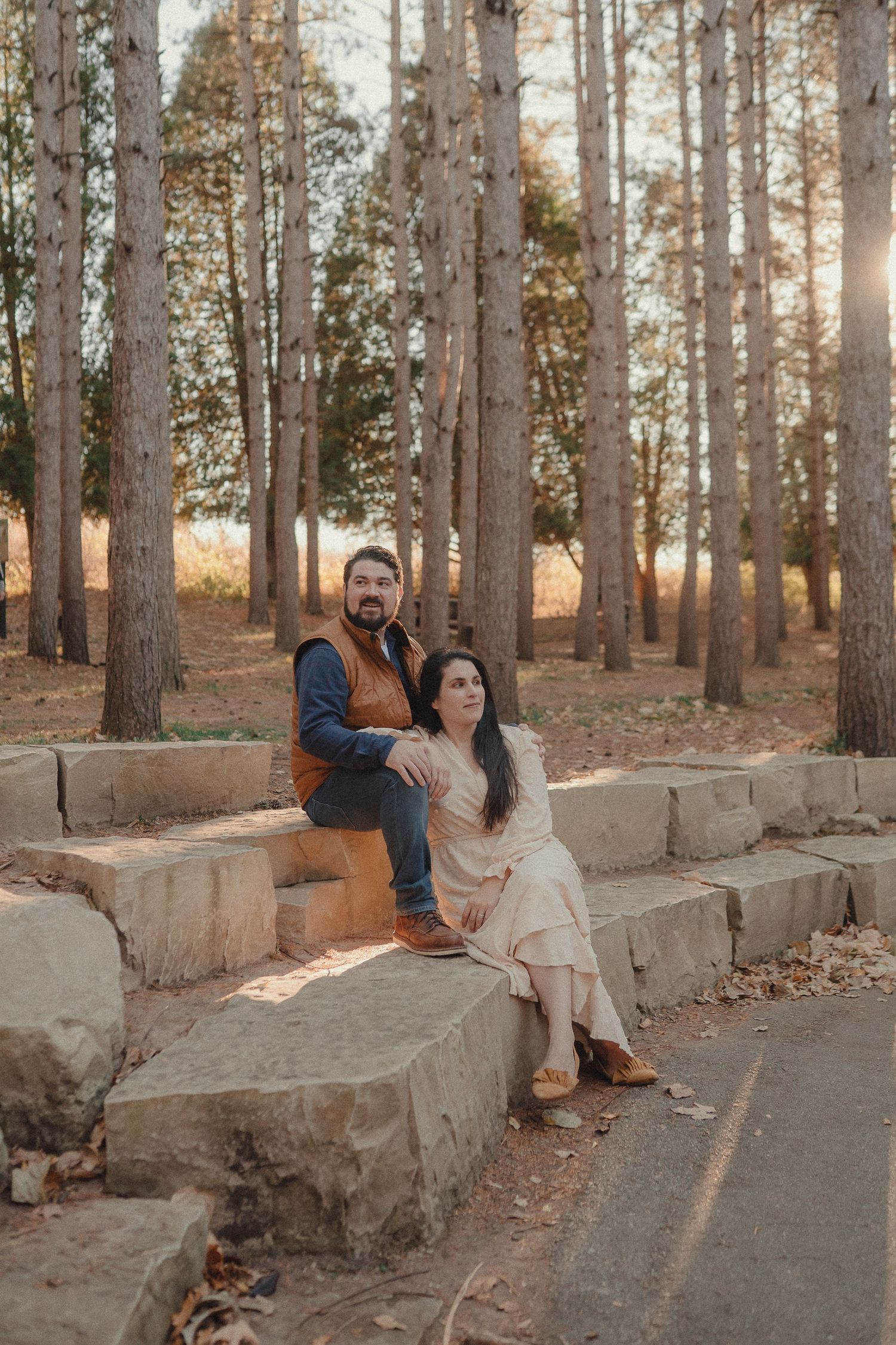 A couple sits together on a stone wall in a sunlit forest path during golden hour.