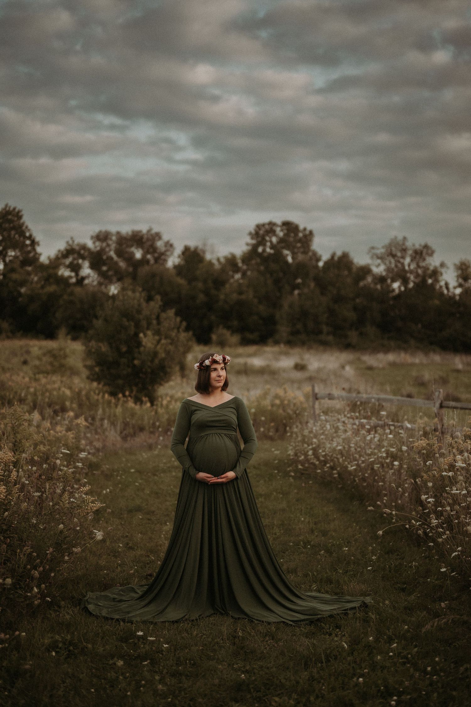 A pregnant woman in a flowing olive green dress stands in a wildflower field at dusk.