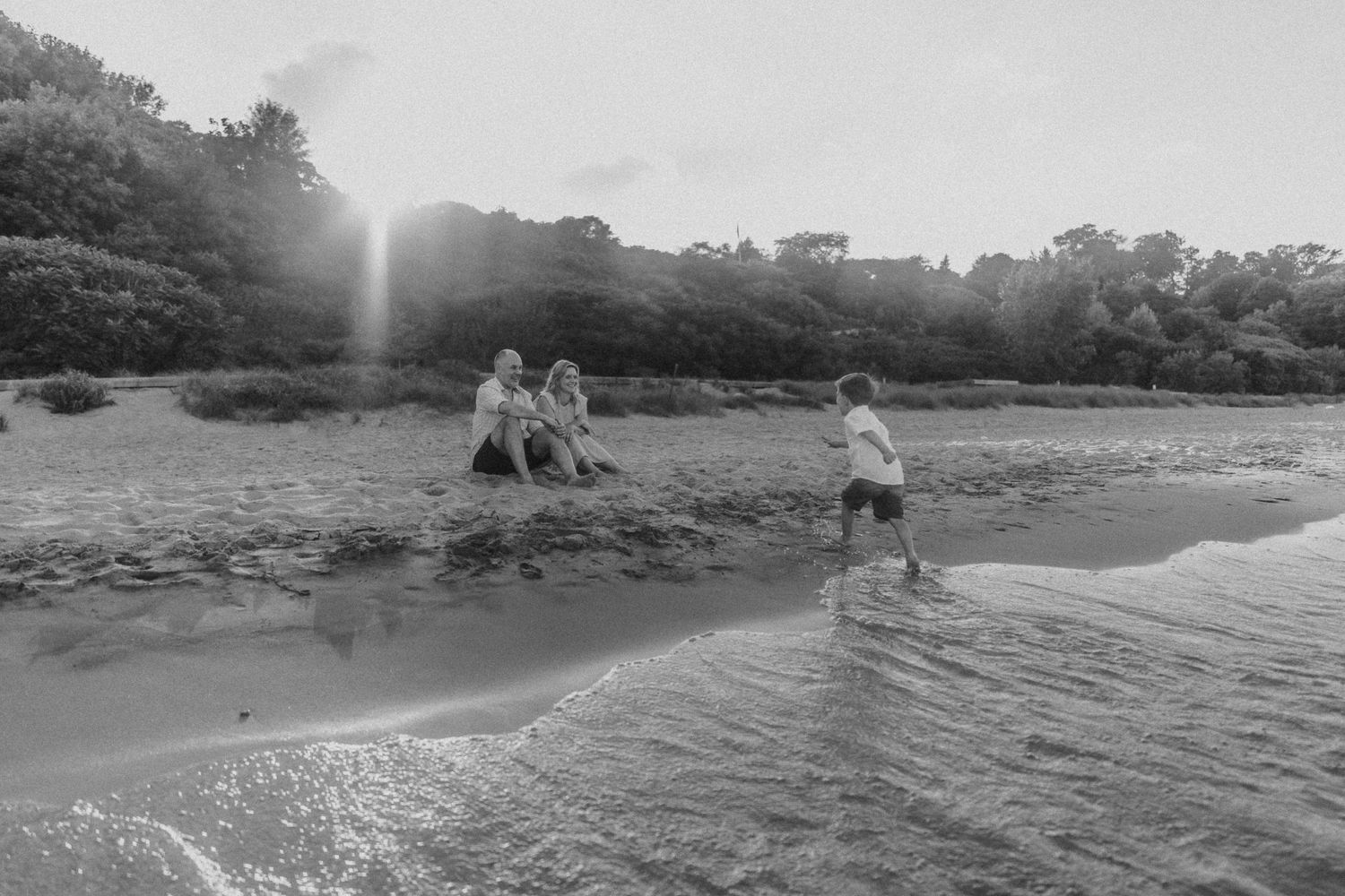 Black and white photo of beachgoers playing in the surf near a wooded coastline.