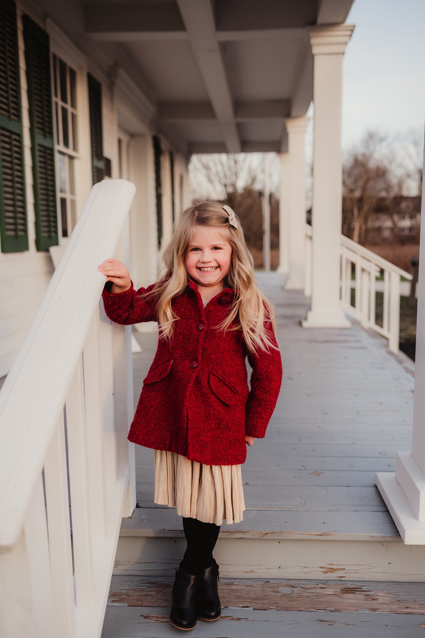 A small child poses on a porch wearing a red coat, pleated skirt and black boots against white columns.