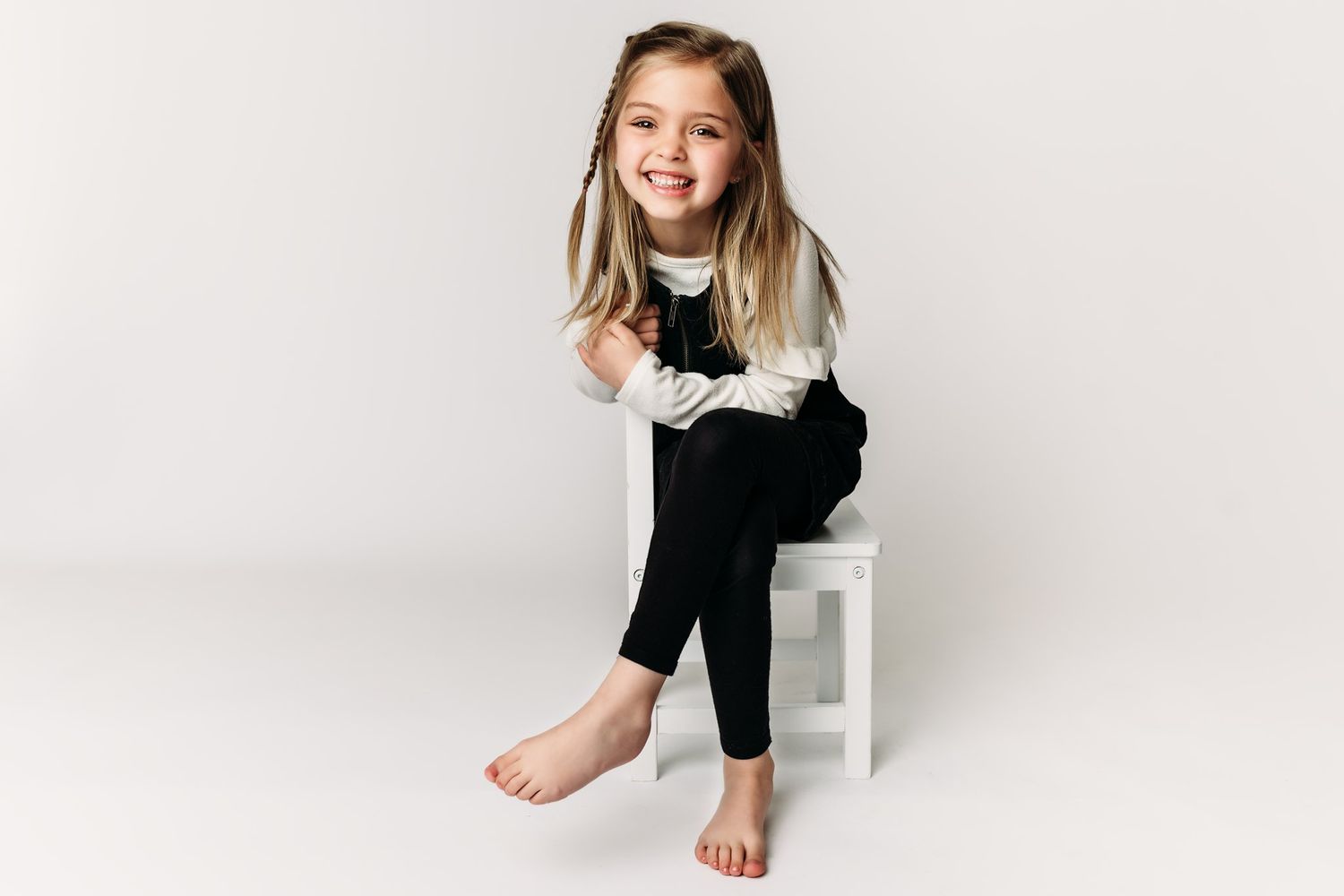 A child sits on a white stool wearing a black and white outfit while smiling.